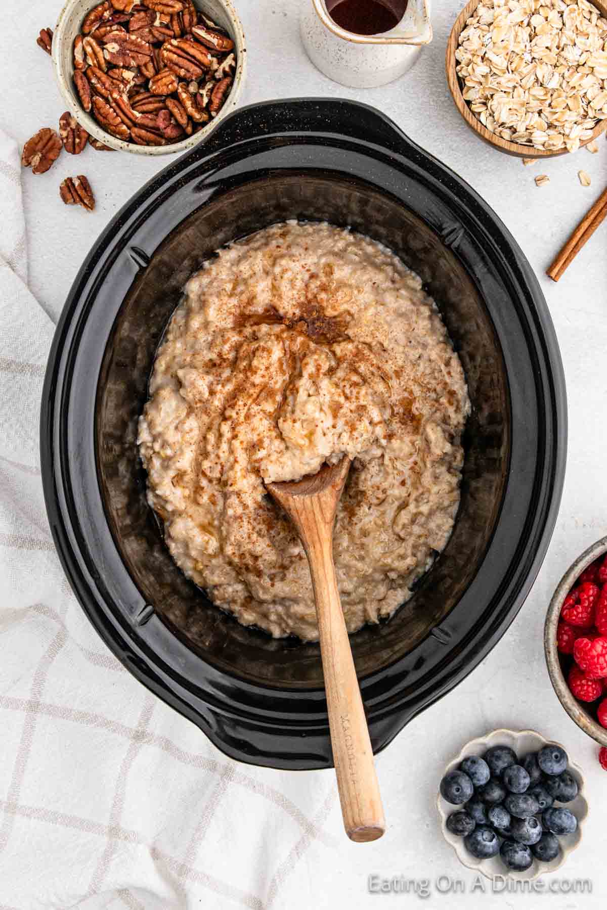 A Crock Pot filled with oatmeal topped with a sprinkle of cinnamon and a wooden spoon. Surrounding the slow cooker are small bowls containing pecans, maple syrup, oats, raspberries, and blueberries, all on a white tablecloth.