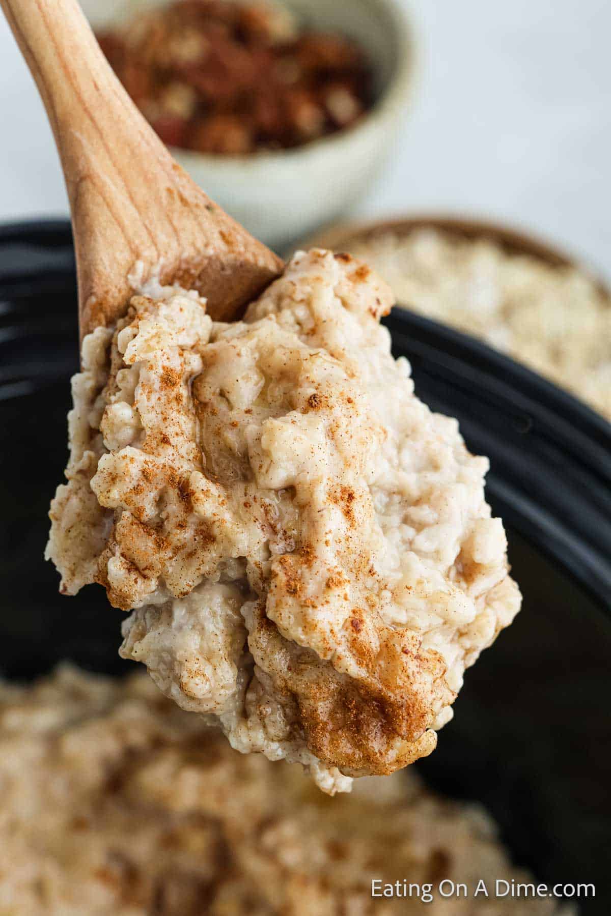 A wooden spoon holds a portion of creamy, cinnamon-dusted oatmeal above a black Crock Pot. In the background, there are two blurred bowls with more oatmeal and nuts. The text "Eating On A Dime.com" is visible in the bottom right corner.