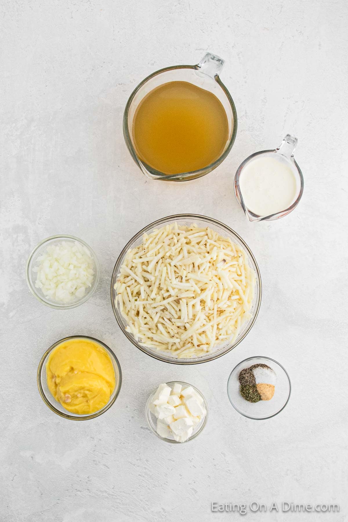 Top-down view of several ingredients for potato soup, including shredded potatoes, diced onions, shredded cheese, broth, milk, seasonings, and cream cheese in bowls and measuring cups on a white surface. A watermark at the bottom right reads "Eating On A Dime.com".