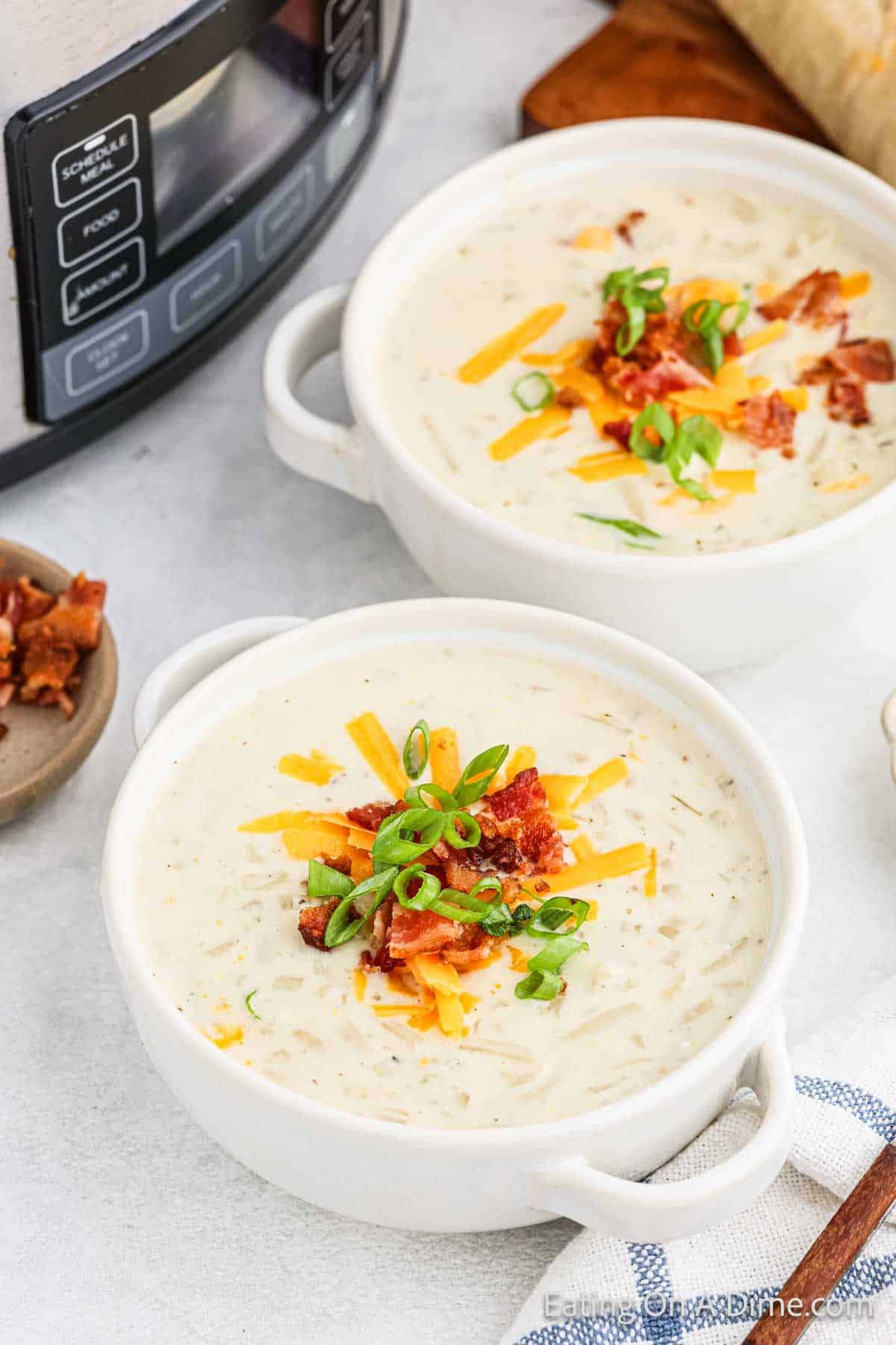 Two white bowls of creamy potato soup, made even heartier with hashbrowns, are garnished with shredded cheddar cheese, crispy bacon bits, and sliced green onions. A Crock Pot is partially visible in the background along with a small bowl of more bacon bits and part of a loaf of bread.