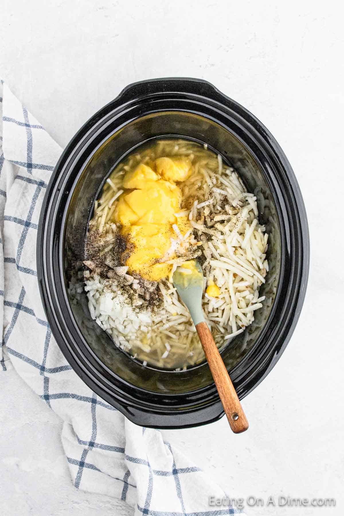 A Crock Pot filled with ingredients, including shredded hashbrowns, melted cheese, seasonings, and chopped onions. A spatula with a wooden handle rests inside the cooker. A checkered cloth is partially visible on the side. Text "Eating On A Dime.com" appears on the bottom right.
