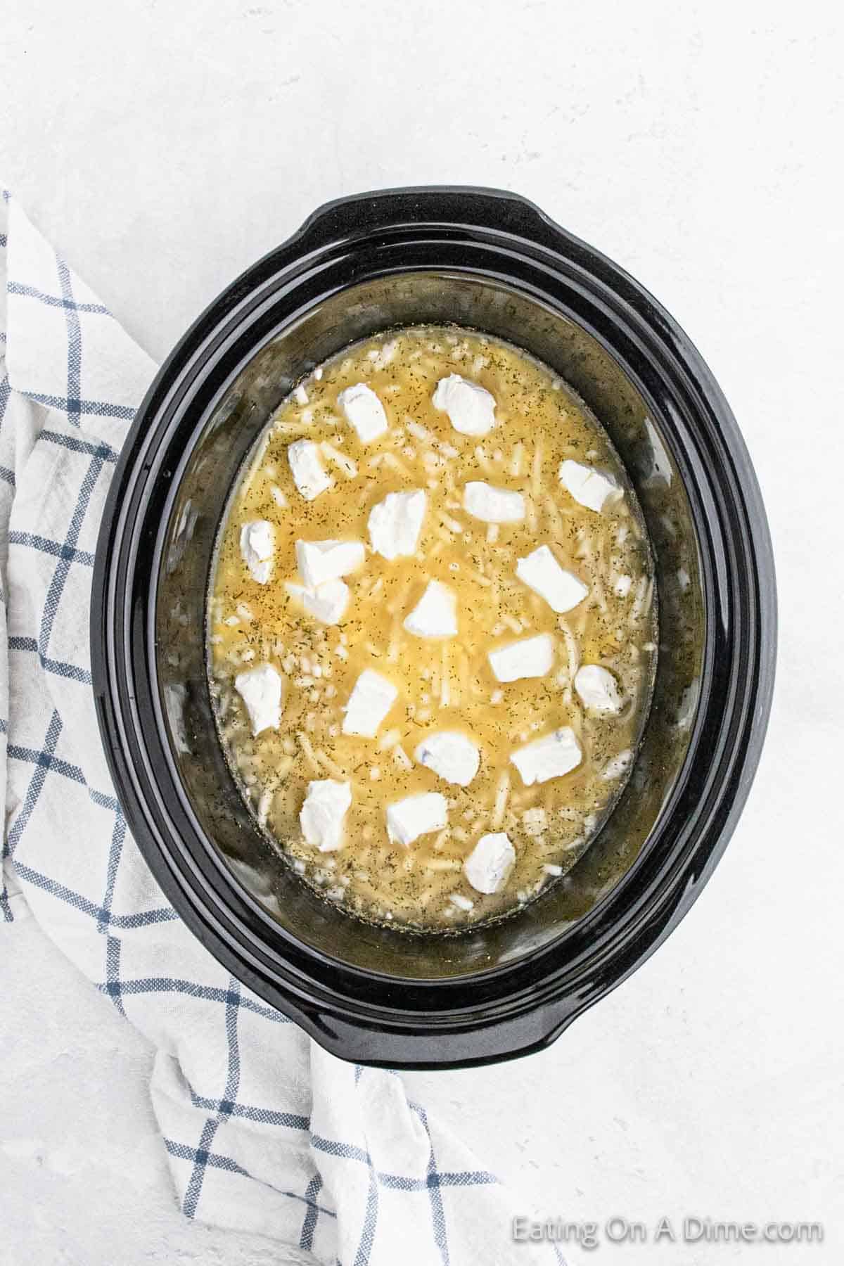 A black Crock Pot filled with a yellowish mixture topped with white dollops, set on a white surface with a blue and white checkered cloth nearby. The contents appear to be partially melted potato soup with some seasoning visible.