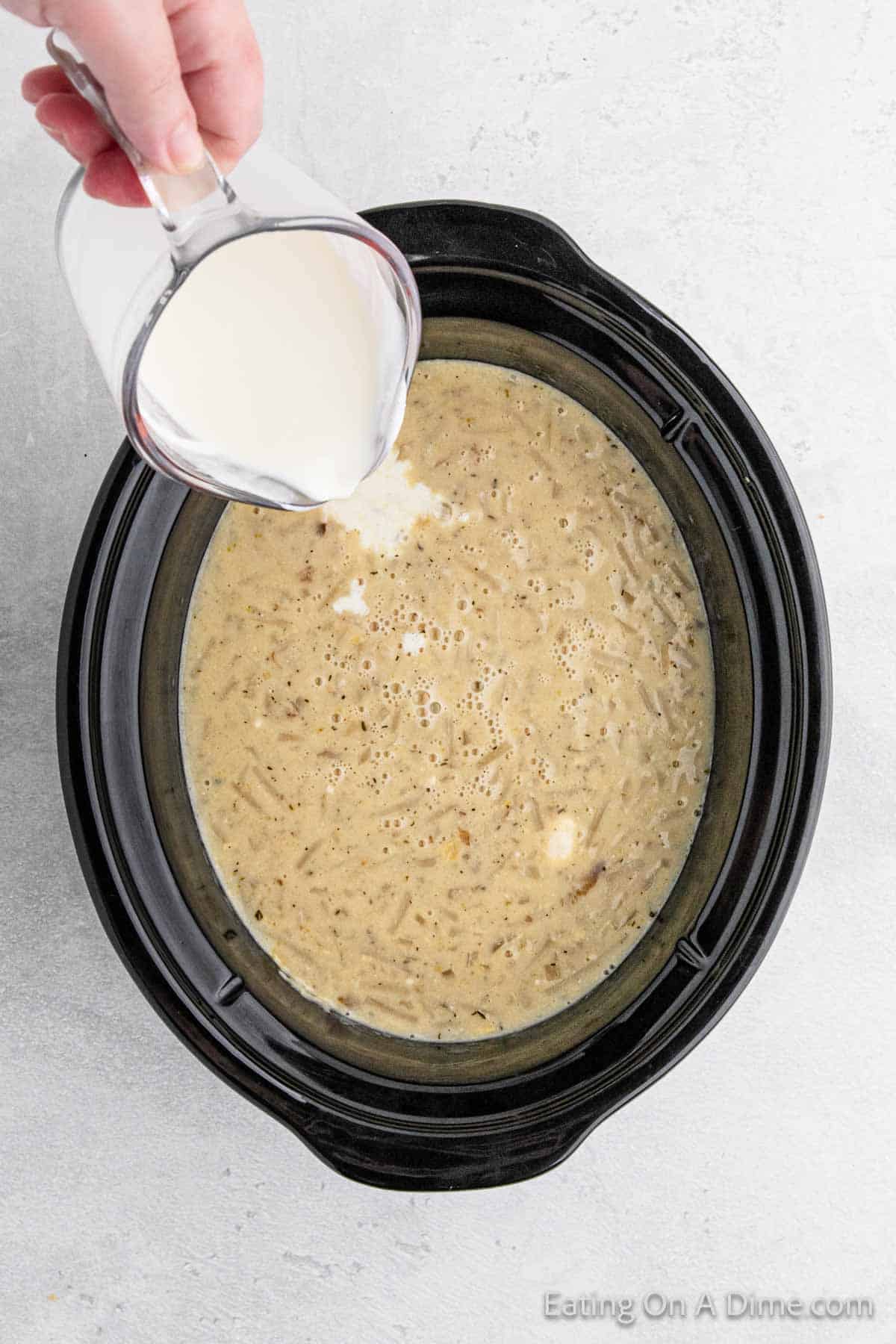 A hand pours cream from a small pitcher into a Crock Pot filled with creamy, seasoned potato soup. The slow cooker is black, and the background is a light, textured surface. The image is labeled with the text "Eating On A Dime.com" in the lower right corner.