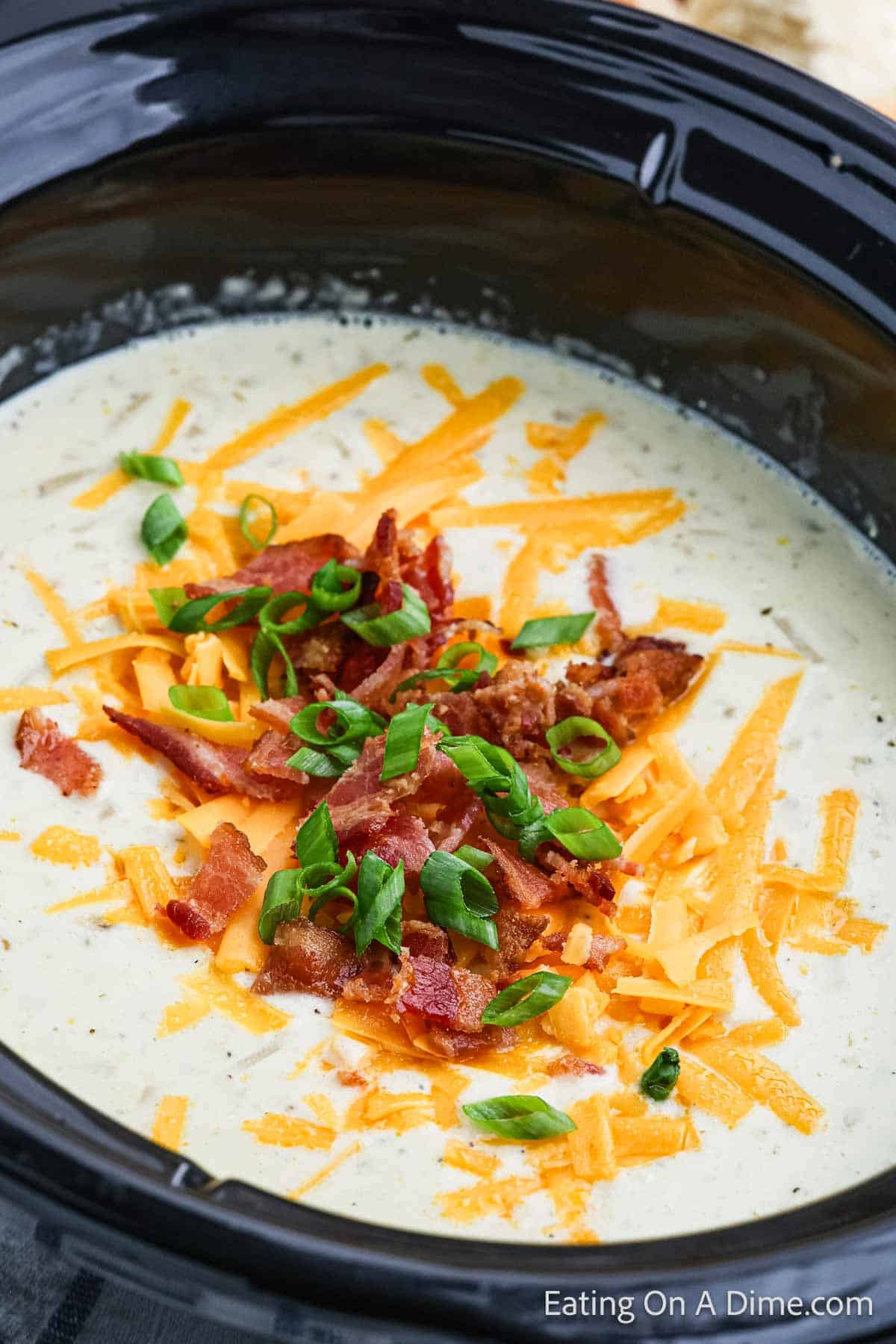 A bowl of creamy potato soup topped with shredded cheddar cheese, crumbled bacon, and chopped green onions. The crock pot soup is served in a black dish and garnished for a colorful and appetizing presentation. The image has a watermark that reads "Eating On A Dime.com".