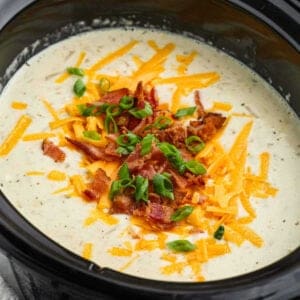 A creamy potato soup in a slow cooker is topped with shredded cheddar cheese, crispy bacon bits, and sliced green onions. The rich and hearty crock pot creation features specks of pepper visible throughout. The toppings add a colorful and appetizing contrast to the dish.