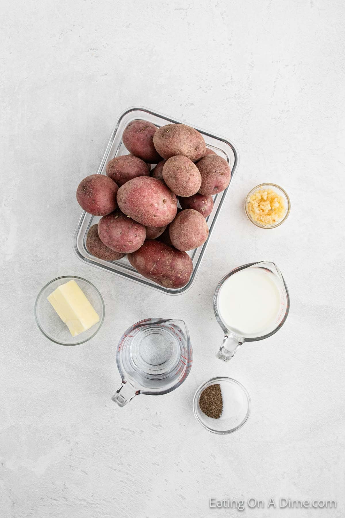 A collection of ingredients for Mashed Red Potatoes: a glass dish filled with red potatoes, a bowl of minced garlic, a measured amount of butter, a jug of milk, a bowl of water, and a small dish of pepper on a light background.