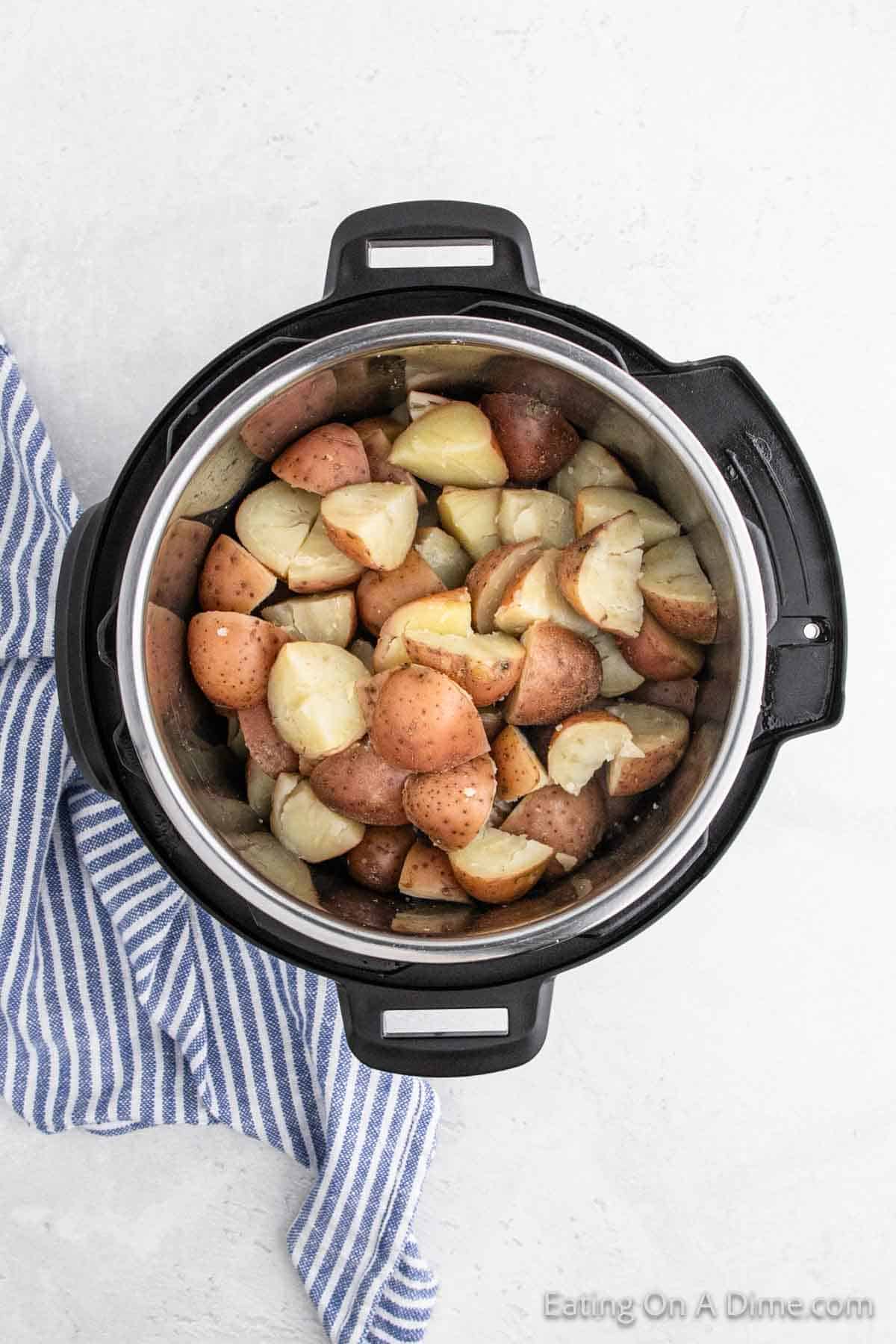 An Instant Pot filled with quartered red potatoes awaits transformation into creamy mashed red potatoes. A blue and white striped cloth is partially visible next to the cooker on a light surface, hinting at the cozy kitchen setting.