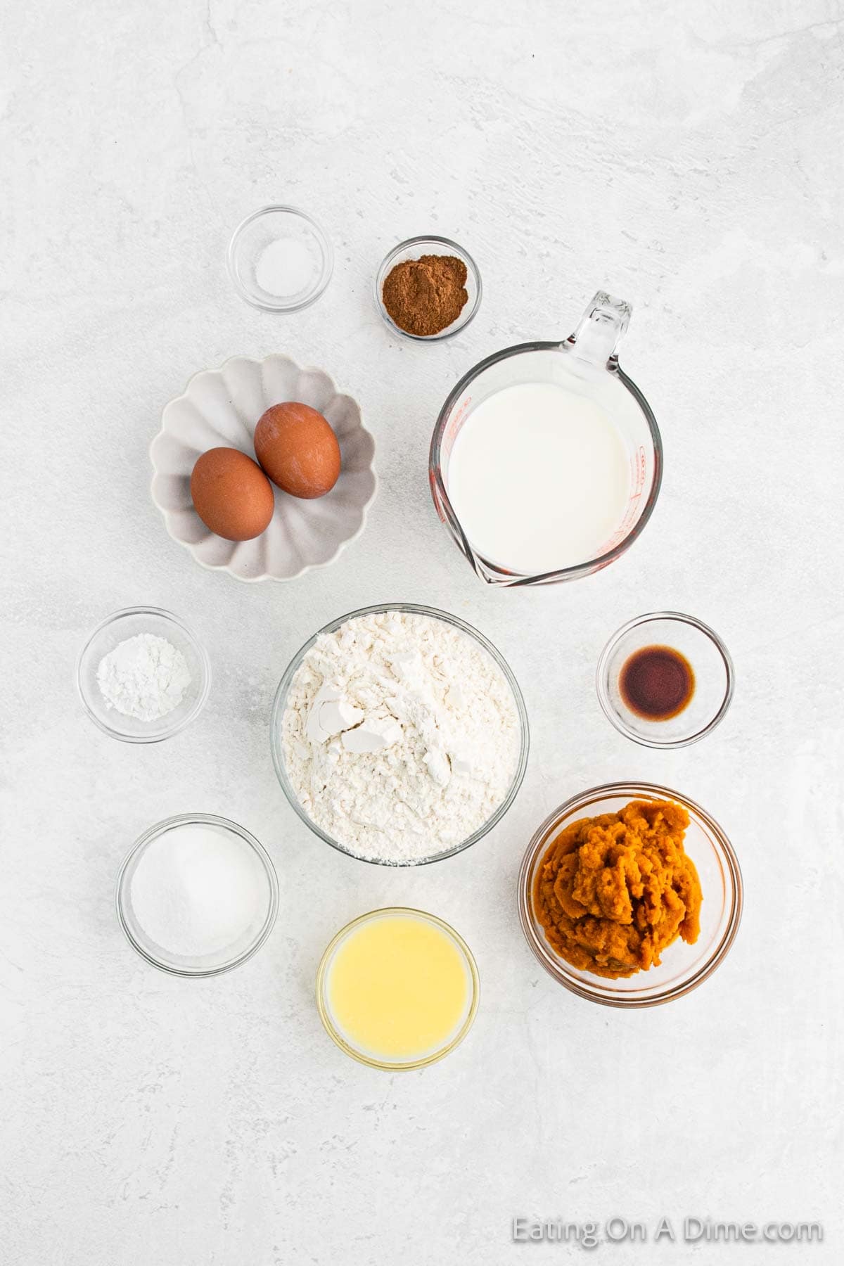 A top-down view of baking ingredients on a white surface. The items include two eggs, a measuring cup of milk, bowls of flour, sugar, pumpkin puree for the waffles recipe, melted butter, vanilla extract, baking powder, cinnamon, and a small bowl of salt.