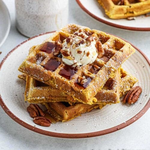 A stack of pumpkin waffles topped with a dollop of whipped cream, maple syrup, and chopped pecans. The waffles are dusted with powdered sugar and served on a white plate with a brown rim. A white pitcher is visible in the background, completing the autumn-inspired breakfast scene.