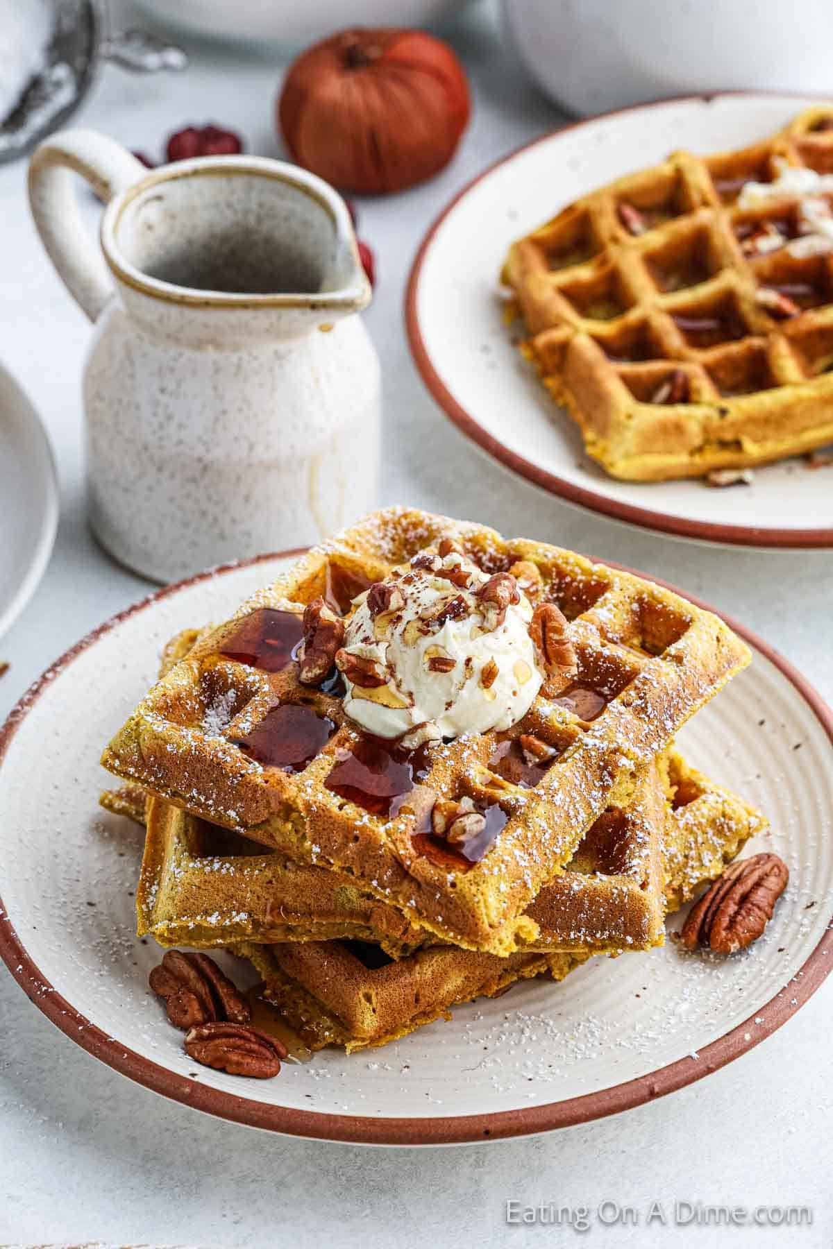 A stack of pumpkin waffles topped with a scoop of ice cream, syrup, and chopped pecans is served on a white plate. A small jug of syrup sits nearby, with another plate of waffles and a small pumpkin in the background. The words "Eating On A Dime.com" are at the bottom right.