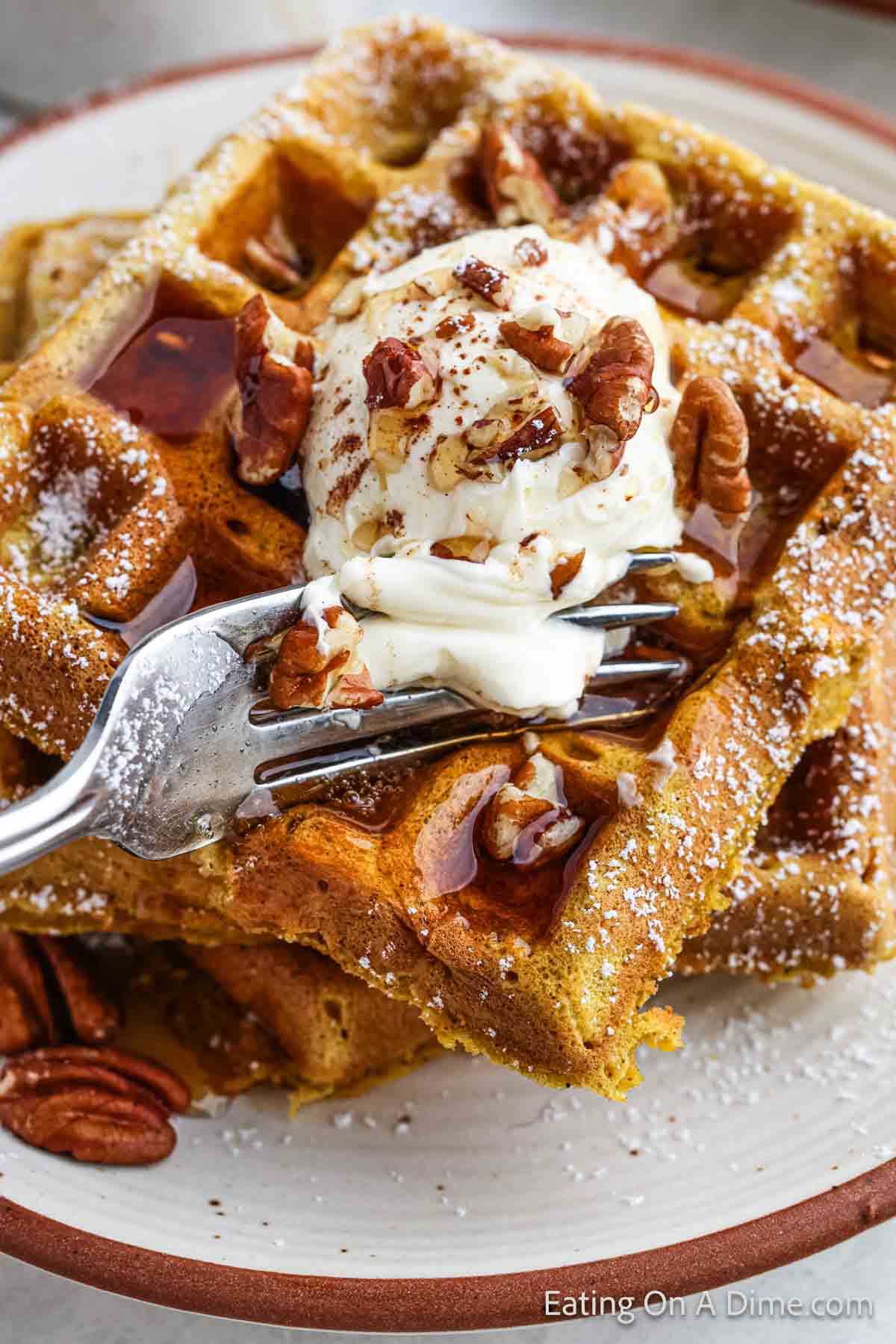 A plate with two Belgian waffles topped with a scoop of melting vanilla ice cream, chopped pecans, and a drizzle of syrup. A fork is cutting into the pumpkin waffle, and some extra pecans are visible on the side of the plate.