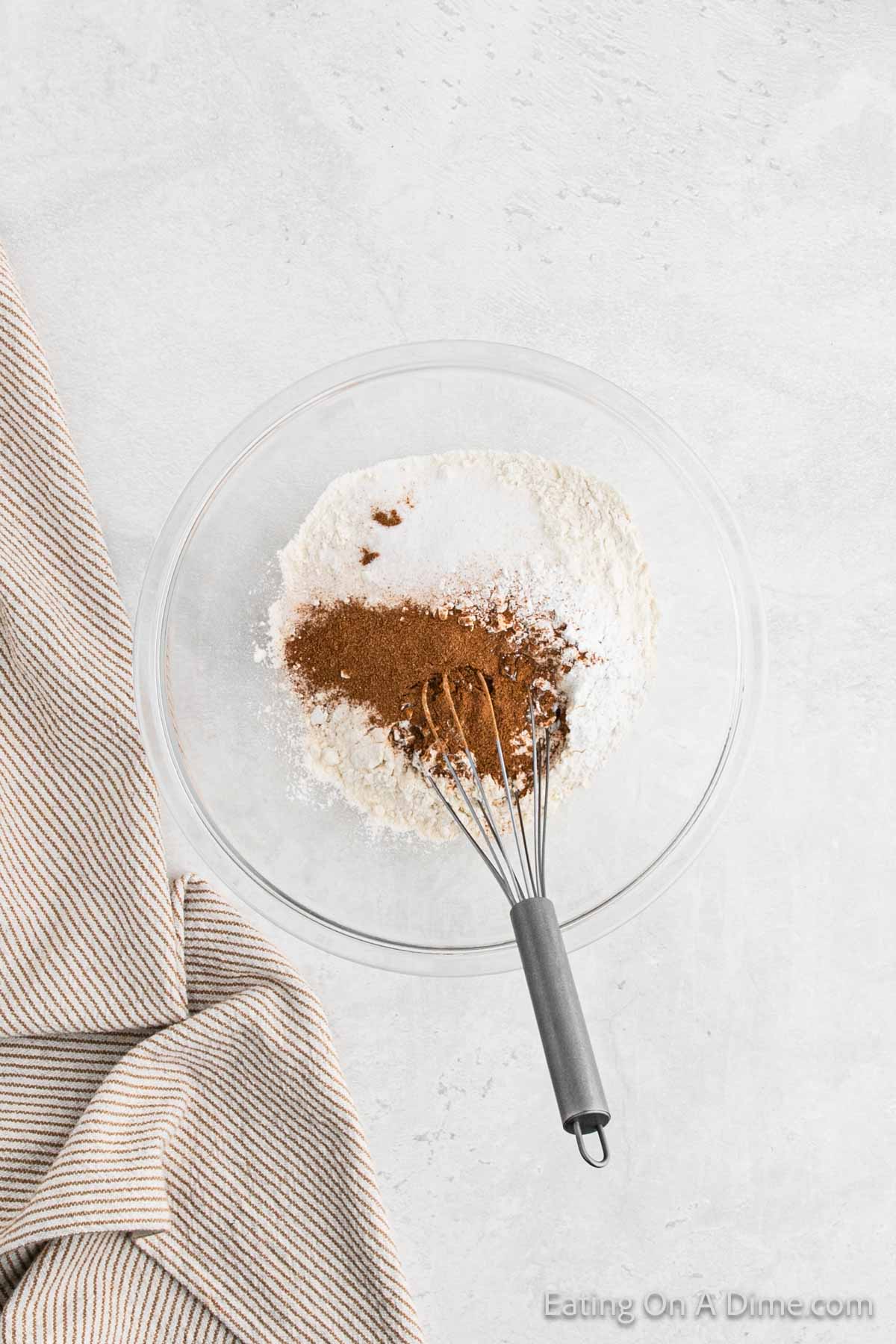 A clear glass mixing bowl on a light surface contains white flour and a mound of brown cinnamon, perfect for making pumpkin waffles. A whisk is partially submerged in the ingredients. A beige and white striped cloth is draped to the left of the bowl. The bottom right has the text "Eating On A Dime.com".