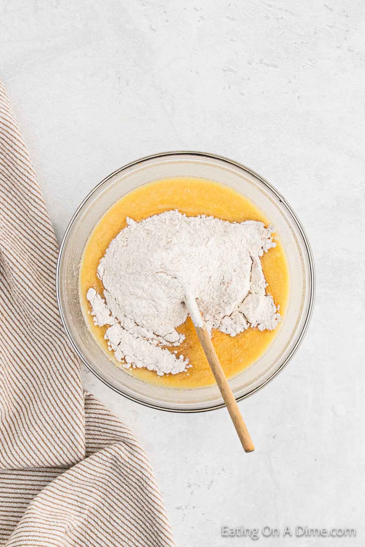 A glass mixing bowl filled with yellow batter for pumpkin waffles and a pile of white flour on top, with a wooden spoon resting inside the bowl. The bowl is placed on a light gray surface next to a beige and white striped cloth.