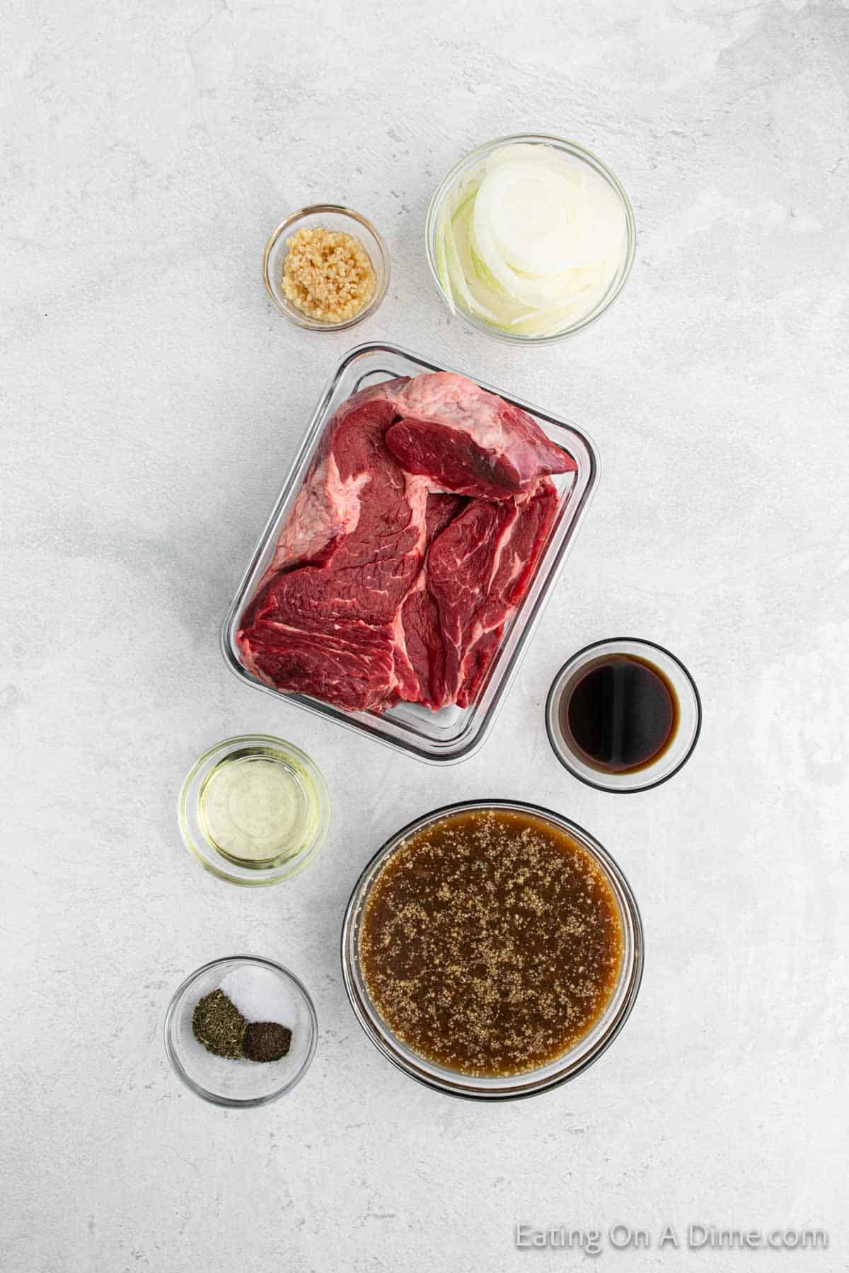 A top-down view of ingredients for a pot roast recipe on a white countertop. Central tray holds raw beef chunks, surrounded by small bowls containing sliced onions, minced garlic, soy sauce, beef broth, seasoned sauce, oil, and green herbs—perfect for your next slow cooker meal.