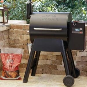 A black Traeger pellet grill stands against a brick background in an outdoor setting, perfect for upcoming Labor Day Sales. A digital control panel is visible on the right, and a bag of apple wood pellets rests beside it. Green foliage can be seen in the background.