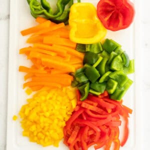 A white cutting board displays a variety of chopped bell peppers. From top to bottom, there are yellow, green, and red peppers sliced into different shapes, including strips and small cubes. Learn how to cut a bell pepper perfectly for any recipe with these diverse cuts.