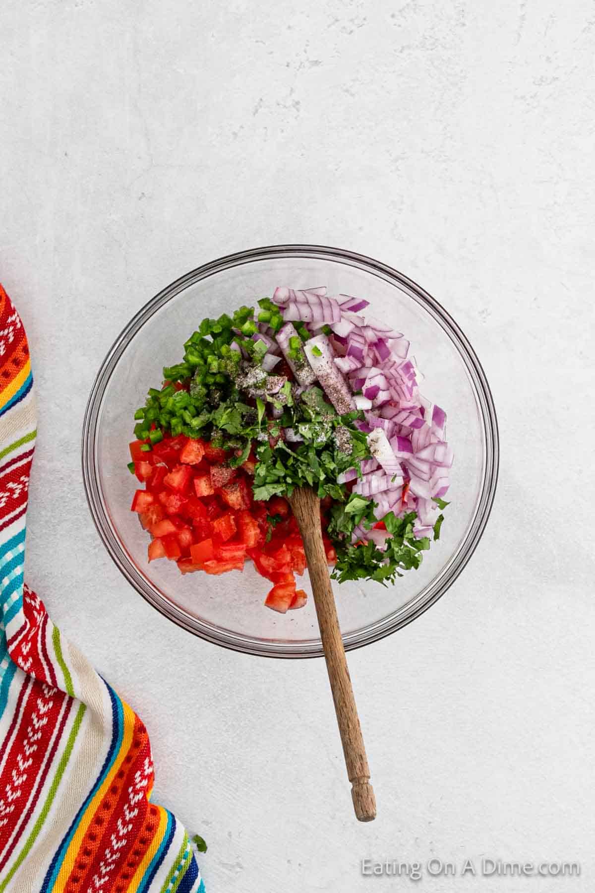 Diced red onions, diced tomatoes, fresh cilantro, and jalapenos in a bowl with a wooden spoon