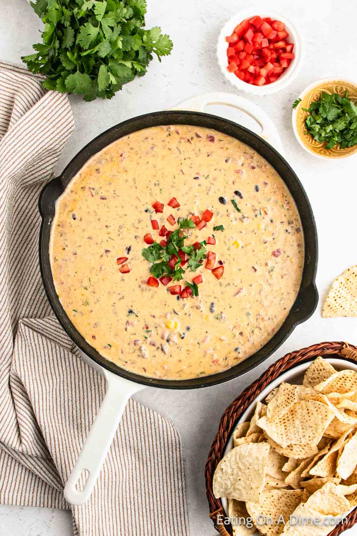 Cowboy queso in a cast iron skillet with small bowls of chopped cilantro, diced tomatoes and tortilla chips