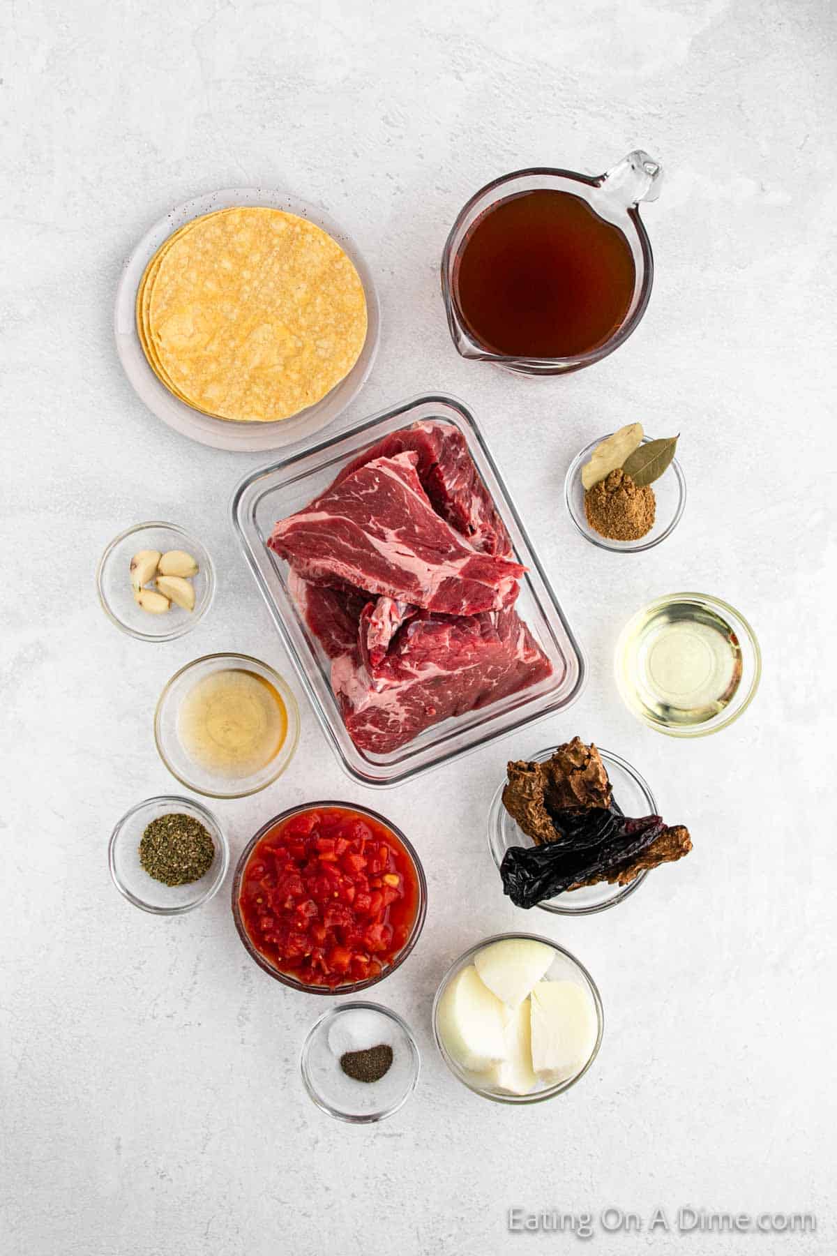 A top-down view of ingredients for a birria recipe, including raw beef cuts, diced tomatoes, garlic cloves, onion, oil, dried chili peppers, tortillas, broth, bay leaf, and various spices in small bowls—all set on a white surface. Perfect for making delicious birria tacos in your Crock Pot.