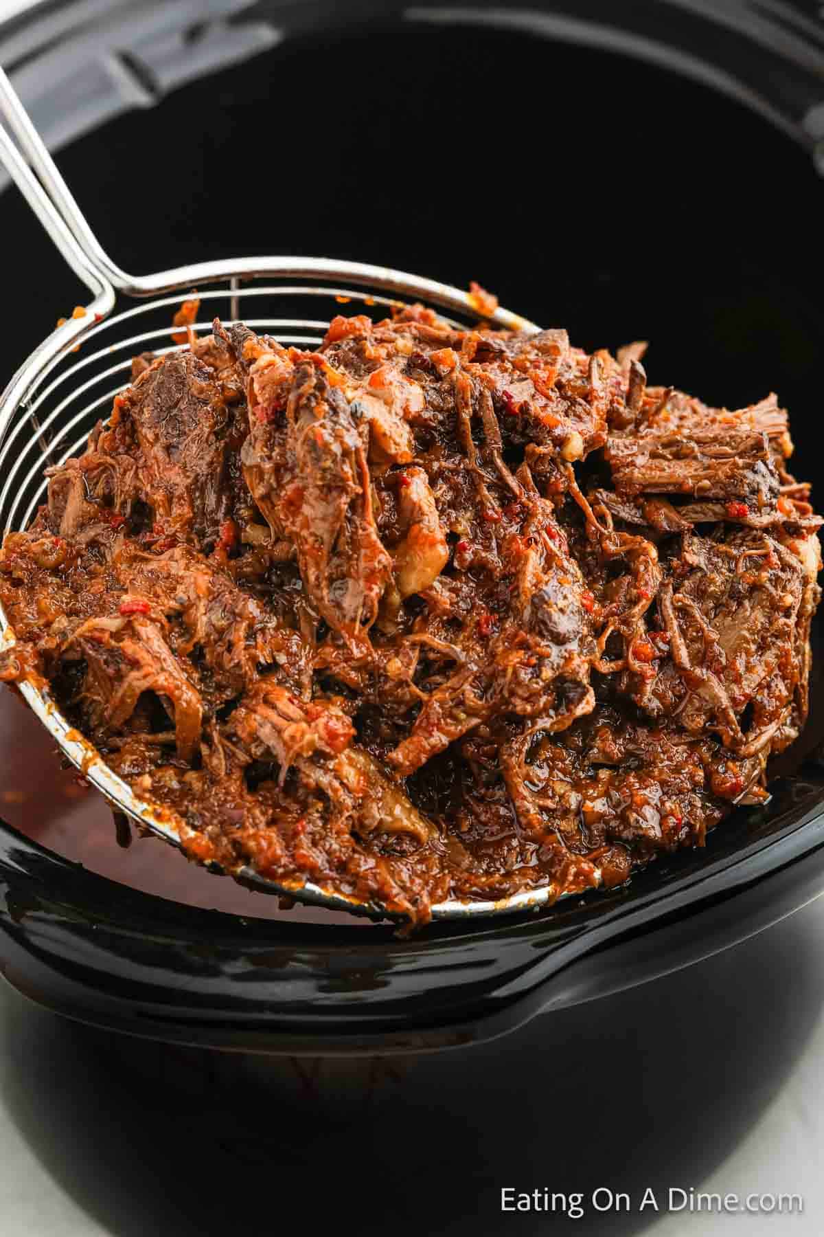 A close-up of a portion of shredded, seasoned birria beef being lifted out of a Crock Pot with a slotted spoon. The beef is dark, tender, and covered in spices and sauce, showcasing its juicy texture. The background is slightly blurred. Perfect for making mouth-watering tacos!