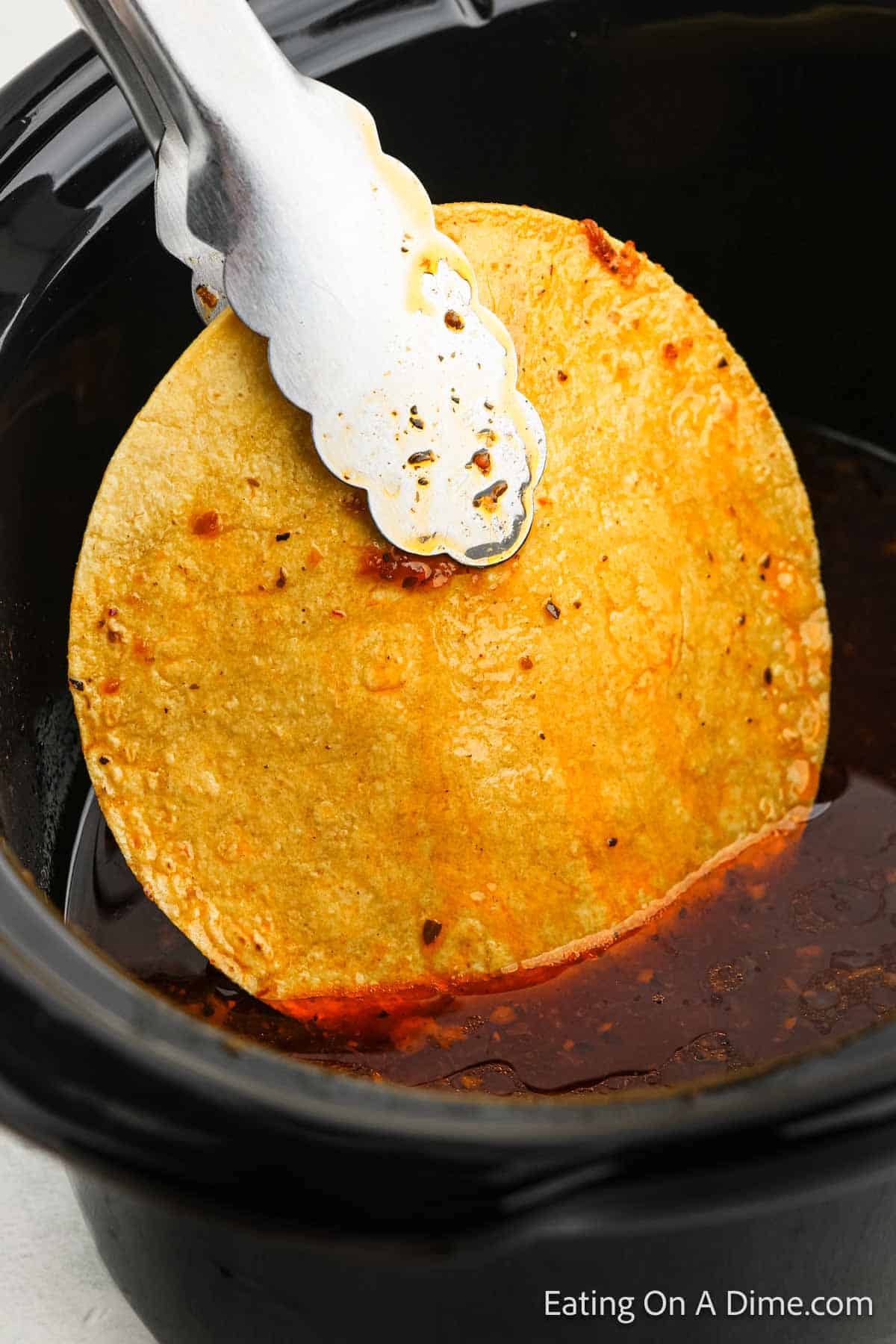 A pair of metal tongs holding a corn tortilla dipped in a rich, reddish-brown sauce inside a black pot. The tortilla is partially submerged, with sauce dripping off the edges, hinting at a flavorful Birria taco being prepared. The text "Eating On A Dime.com" is visible.