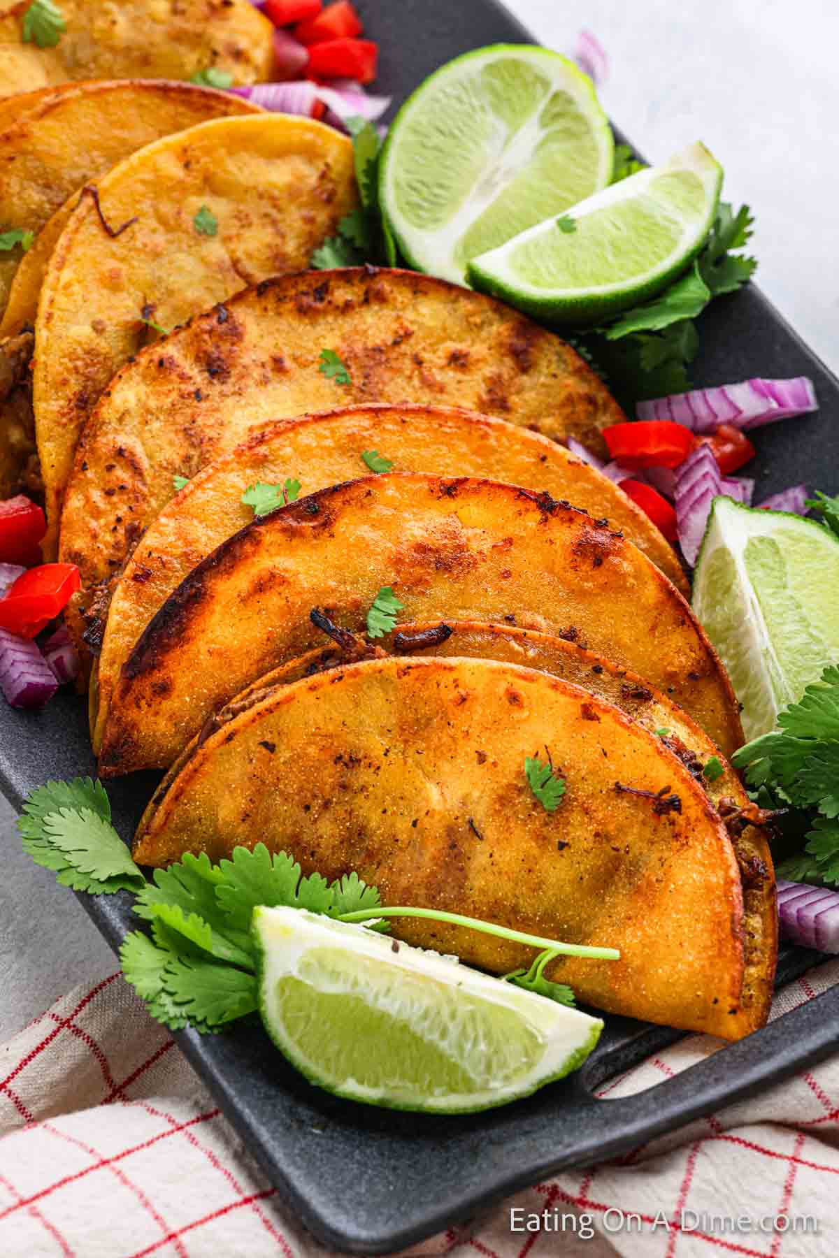 A rectangular black plate with several golden-brown Birria Tacos filled with savory ingredients, garnished with fresh cilantro. Lime wedges, chopped red onions, and pieces of red bell pepper complement the presentation.