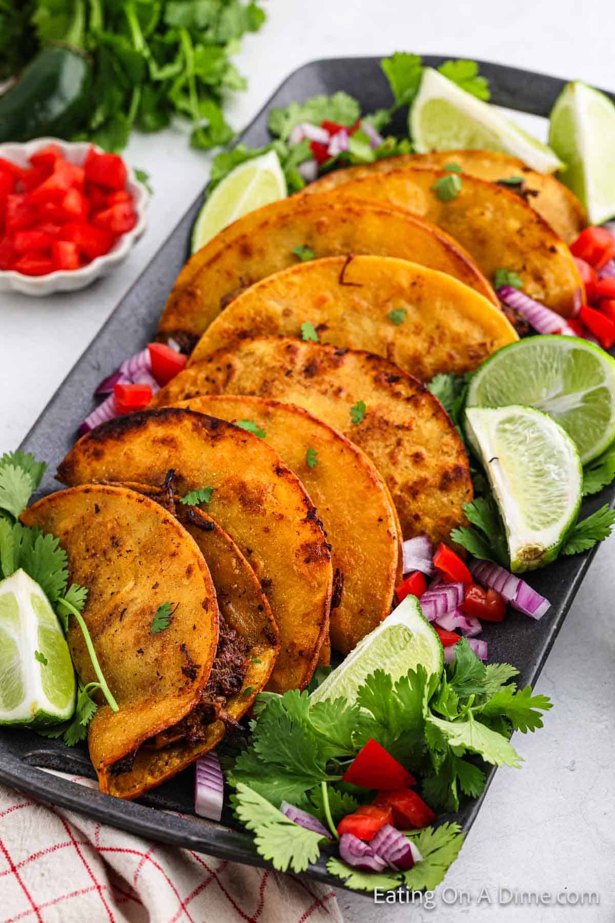 A rectangular black plate contains five crispy Birria Tacos filled with shredded meat made in a Crock Pot, garnished with chopped red onions, cilantro, and lime wedges. The plate is surrounded by additional garnishes including red bell pepper pieces, cilantro leaves, and lime quarters.