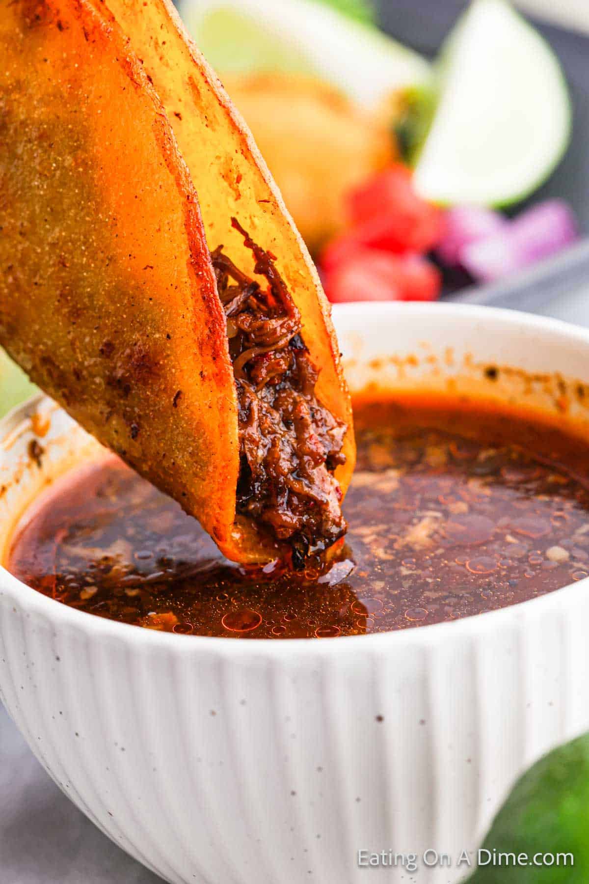 Close-up of a crispy birria taco stuffed with shredded beef being dipped into a bowl of rich, spiced consommé. The taco is partially submerged, showing a glossy coating of the broth. In the background, there are garnishes like lime wedges, chopped onions, and cilantro.