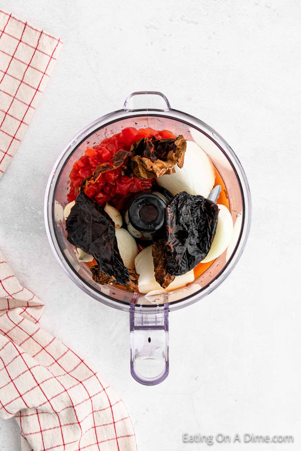 A food processor containing ingredients for a sauce or salsa, perfect for Birria tacos, including chopped tomatoes, onion halves, and dried chili peppers. The processor sits on a light gray surface next to a red and white checkered cloth. "Eating On A Dime.com" is visible in the bottom right corner.