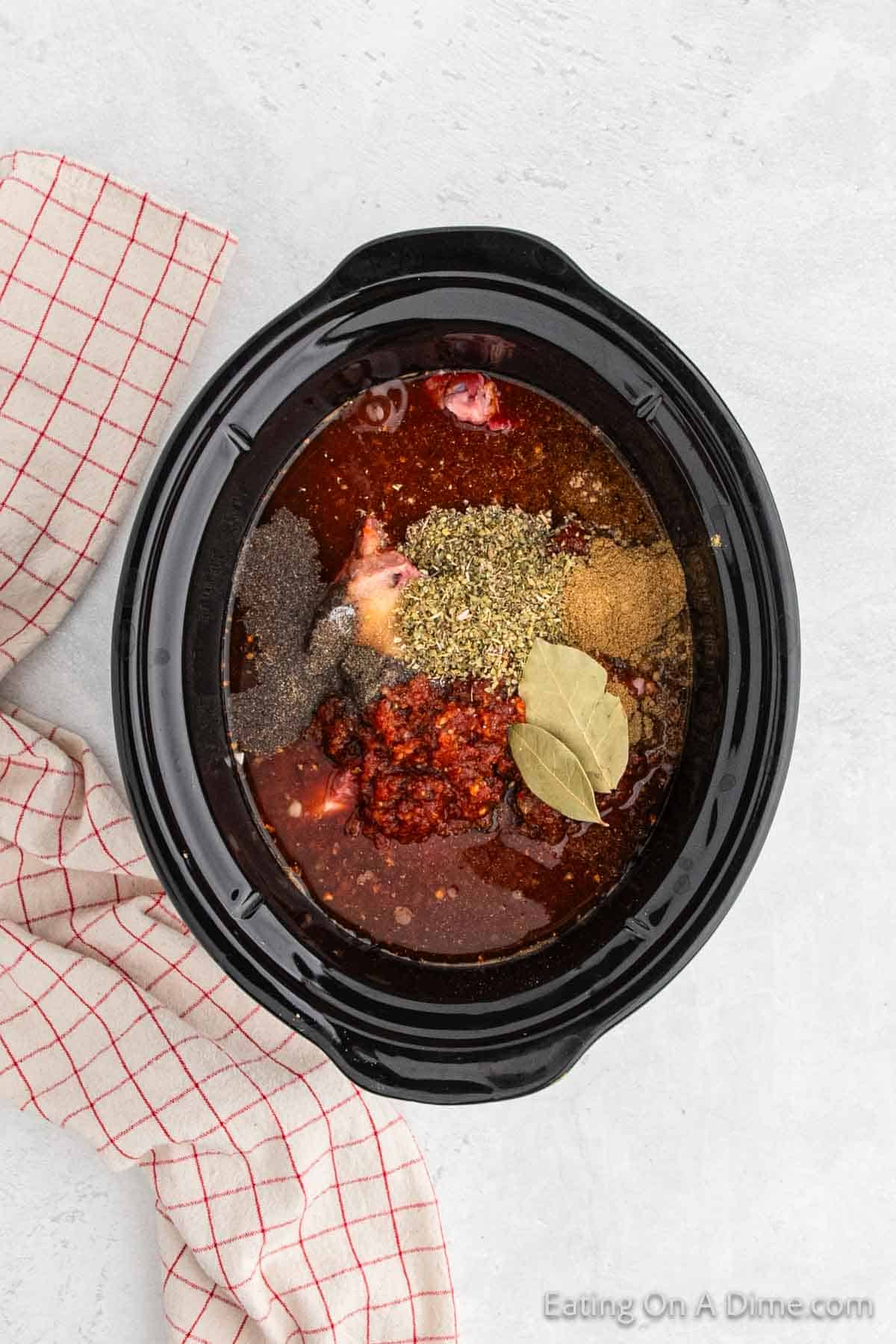 Top view of a black Crock Pot Birria containing raw meat and various seasonings, including ground spices and whole bay leaves. A red and white checkered cloth is partially visible on the left side of the Crock Pot placed on a light gray surface.