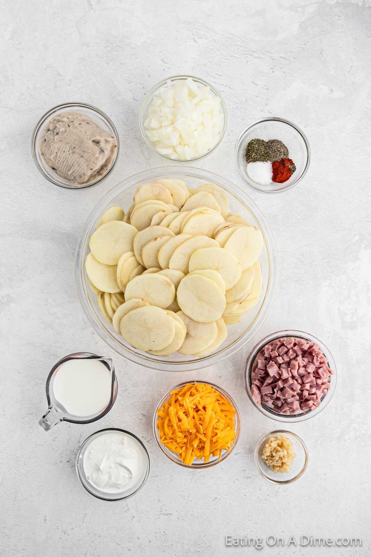 A flat lay of ingredients for Crock Pot Scalloped Potatoes and Ham on a white surface. Items include a bowl of sliced potatoes, a bowl of canned soup, diced onions, grated cheese, diced ham, seasoning, minced garlic, sour cream, and a small pitcher of milk.