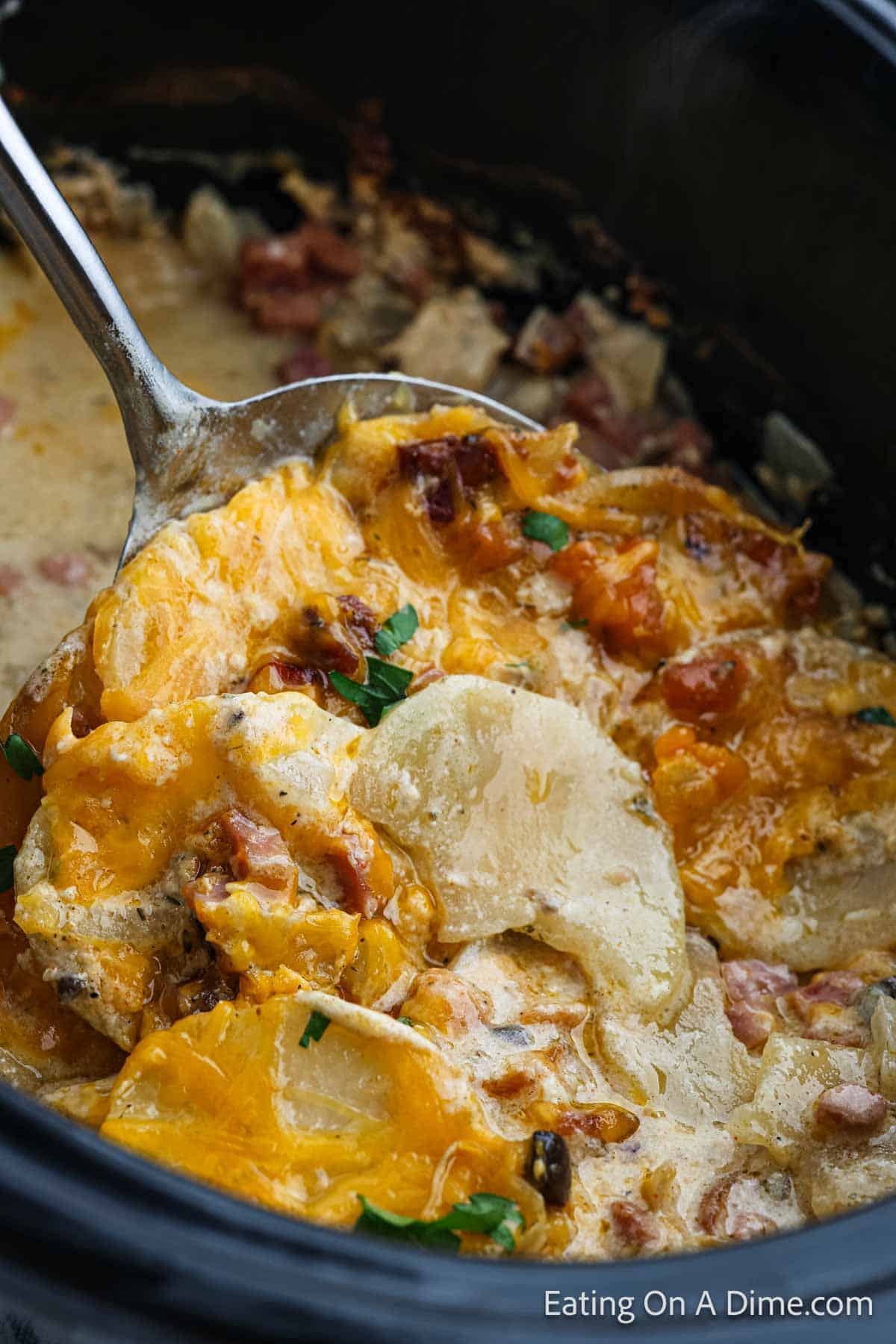 A close-up of a spoon scooping cheesy potatoes and ham from a slow cooker. The dish, reminiscent of Crock Pot Scalloped Potatoes and Ham, is rich in melted cheese, creamy sauce, and garnished with chopped herbs. The background shows more of the creamy, cheesy mixture inside the slow cooker.