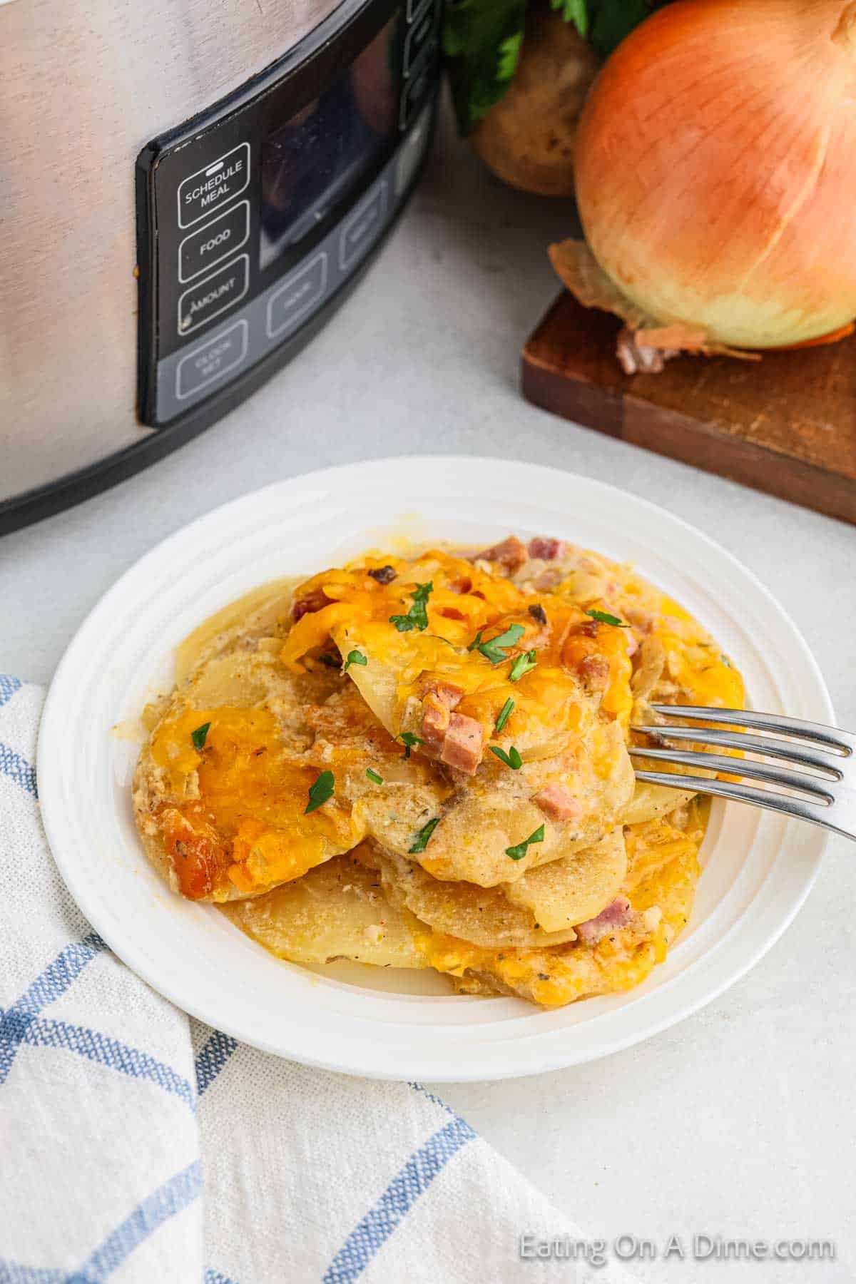 A white plate with a serving of cheesy Crock Pot Scalloped Potatoes and Ham garnished with parsley. The plate is placed near a blue and white checkered napkin. In the background, a slow cooker, an onion, and a wooden cutting board are visible.