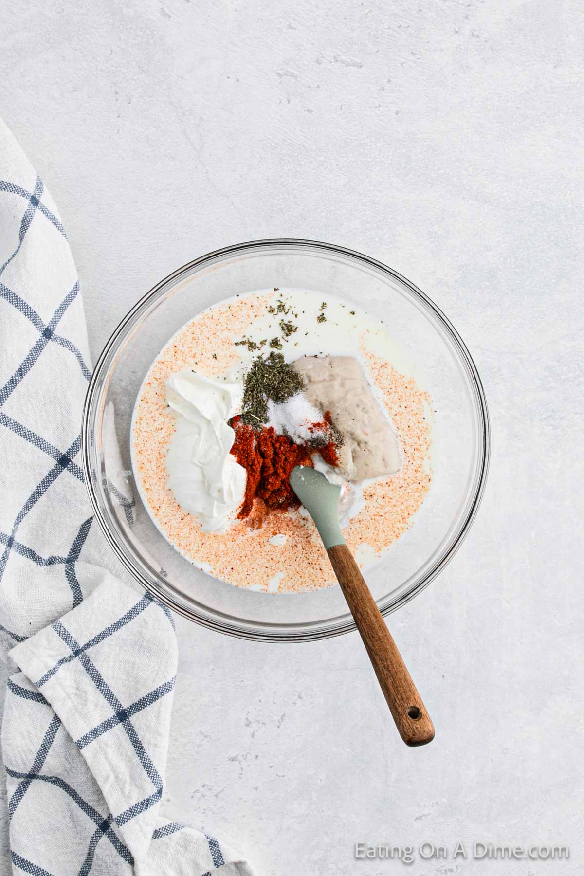 A glass bowl on a white countertop contains a mixture of breadcrumbs, Greek yogurt, red seasoning, and herbs, reminiscent of the flavors found in Crock Pot Scalloped Potatoes and Ham. A gray rubber spatula with a wooden handle rests inside the bowl. A blue and white checkered cloth is partially visible on the left side.