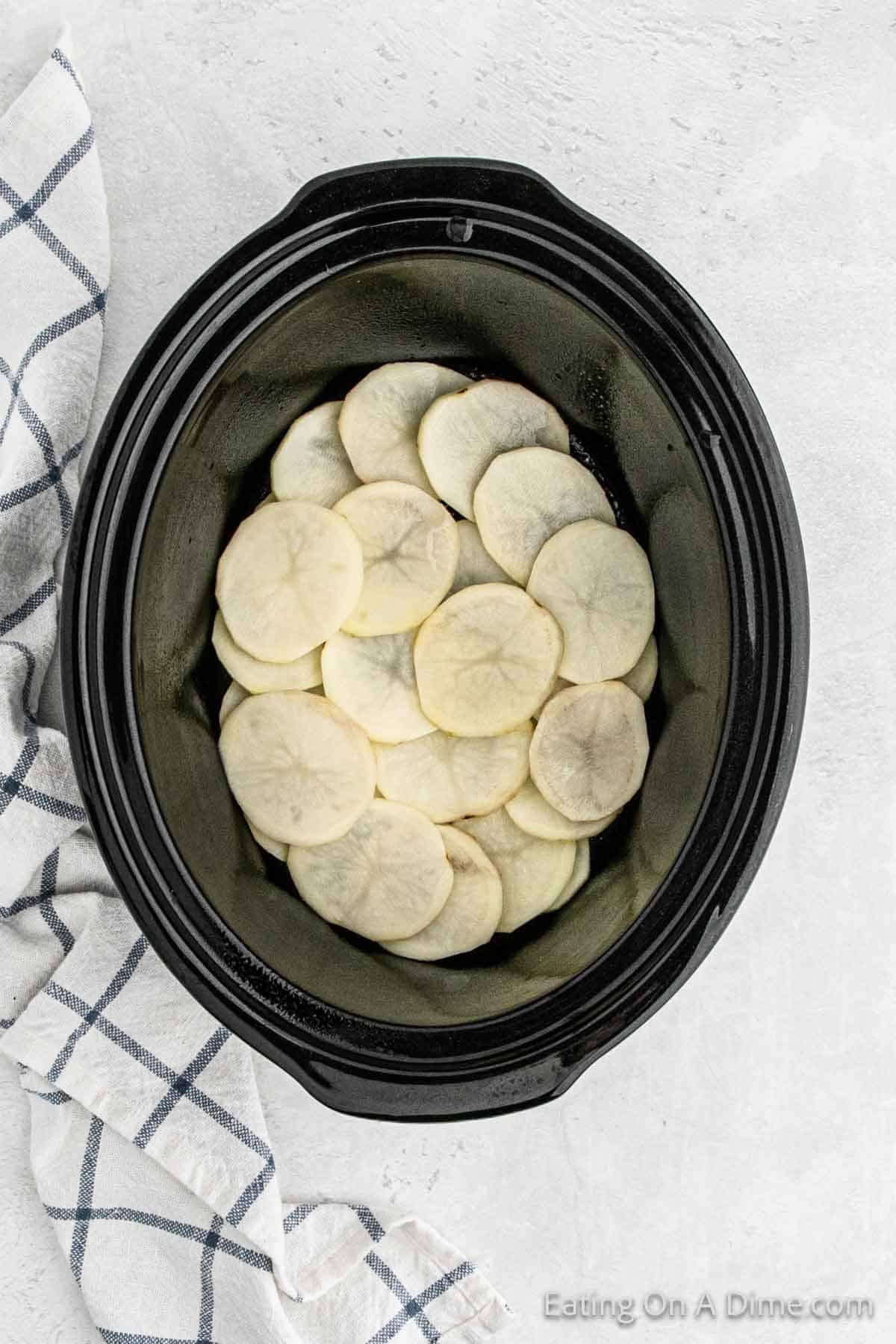 Top view of a black slow cooker with sliced raw potatoes neatly arranged at the bottom. A white cloth with blue checks is placed beside the slow cooker on a light-colored surface. The corner of the image has text reading "Eating On a Dime.com." Perfect for Crock Pot Scalloped Potatoes and Ham.