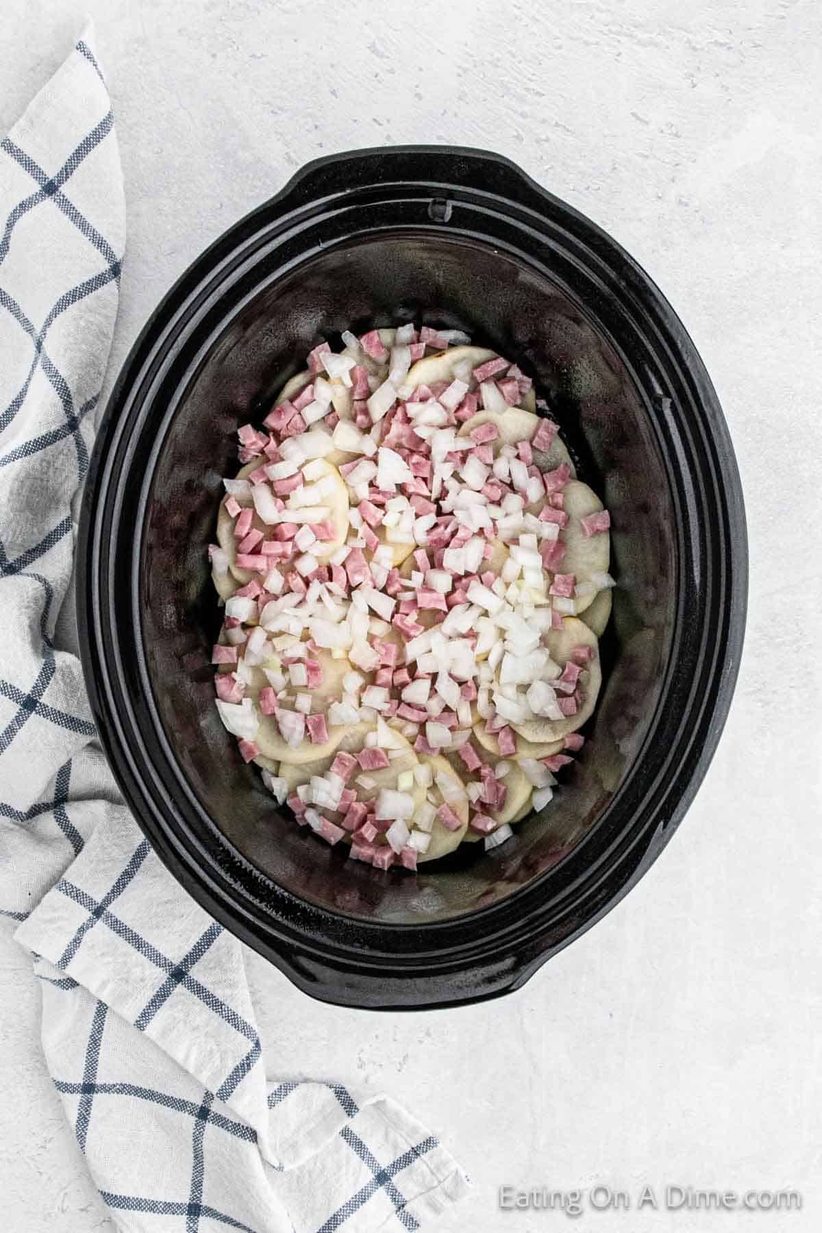 A black slow cooker filled with chopped onions, ham cubes, and sliced potatoes, set on a white countertop. A white kitchen towel with blue grid patterns is draped beside the slow cooker. The “Eating On A Dime” watermark is visible at the bottom right corner. This scene perfectly captures Crock Pot Scalloped Potatoes and Ham.