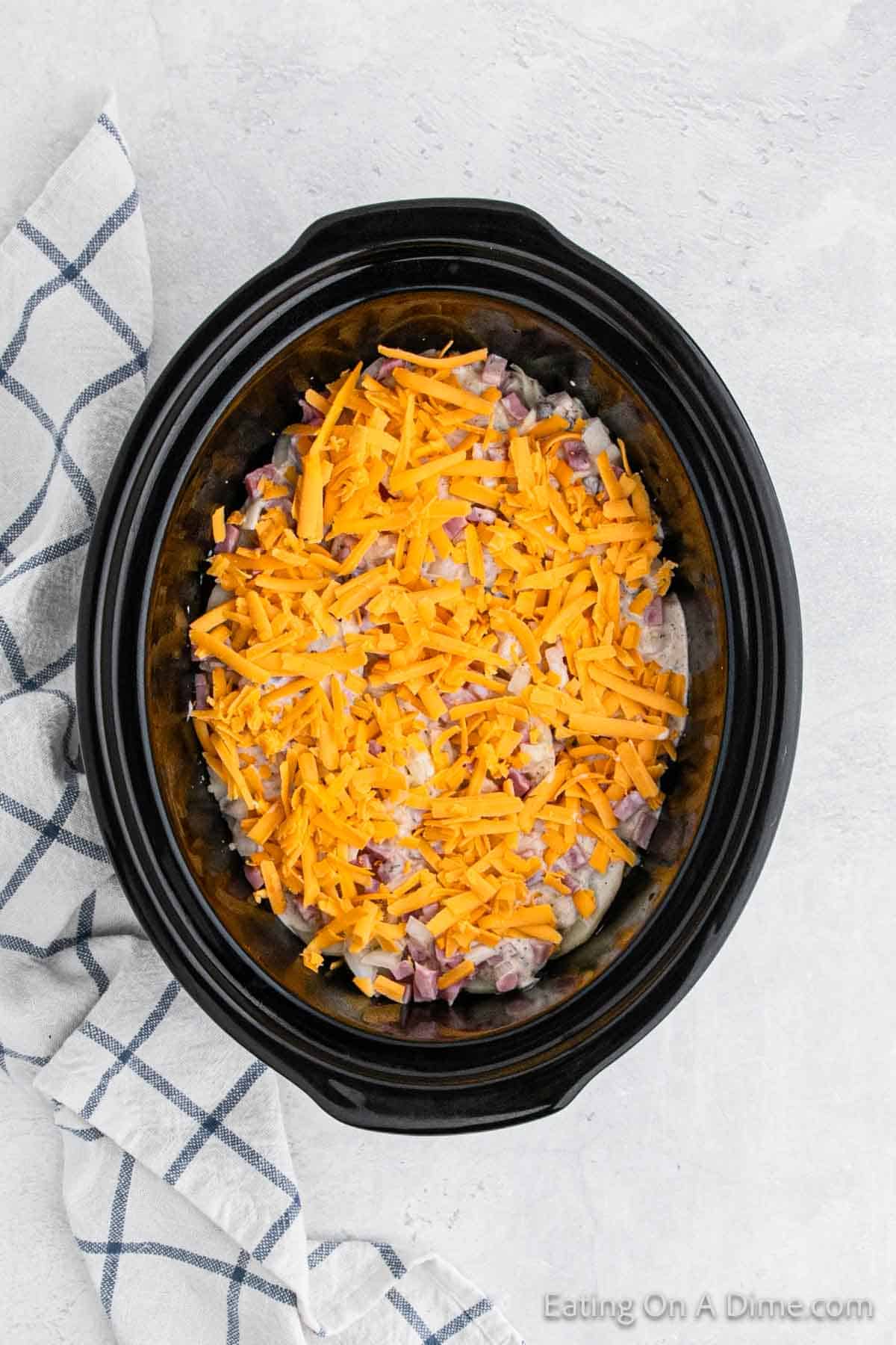 A Crock Pot filled with chopped potatoes, diced onions, and bacon pieces, all topped with a generous layer of shredded cheddar cheese. A white and blue checkered kitchen towel is placed next to the slow cooker on a light grey countertop. Enjoy your Crock Pot Scalloped Potatoes and Ham!