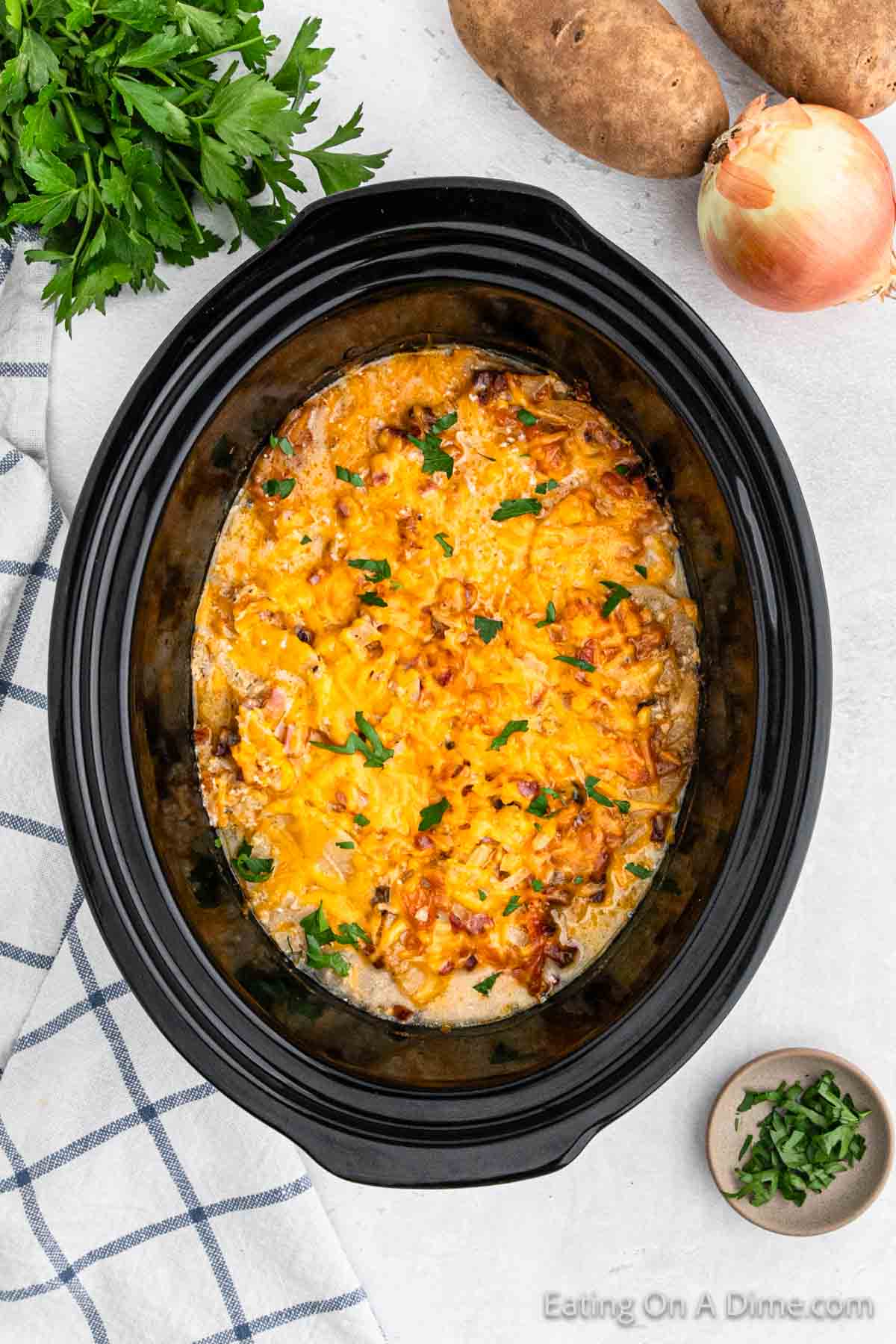 A black slow cooker filled with a cheesy potato casserole topped with melted cheese and chopped parsley. Surrounding the slow cooker are a white and blue checkered cloth, fresh parsley, russet potatoes, and an onion. This delightful dish is perfect for Crock Pot Scalloped Potatoes and Ham lovers.