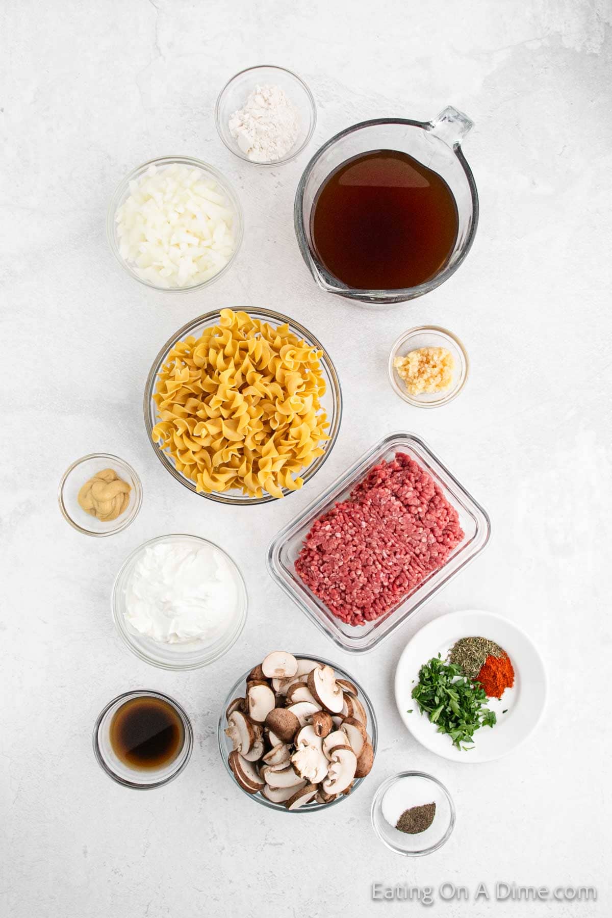 Top-down view of various ingredients spread out on a white surface. Includes bowls of chopped onions, flour, beef broth, egg noodles, minced garlic, ground meat for Stroganoff or Hamburger Helper, mushrooms, sour cream, Worcestershire sauce, mustard, spices, and herbs. "Eating On A Dime.com" is printed at the bottom.