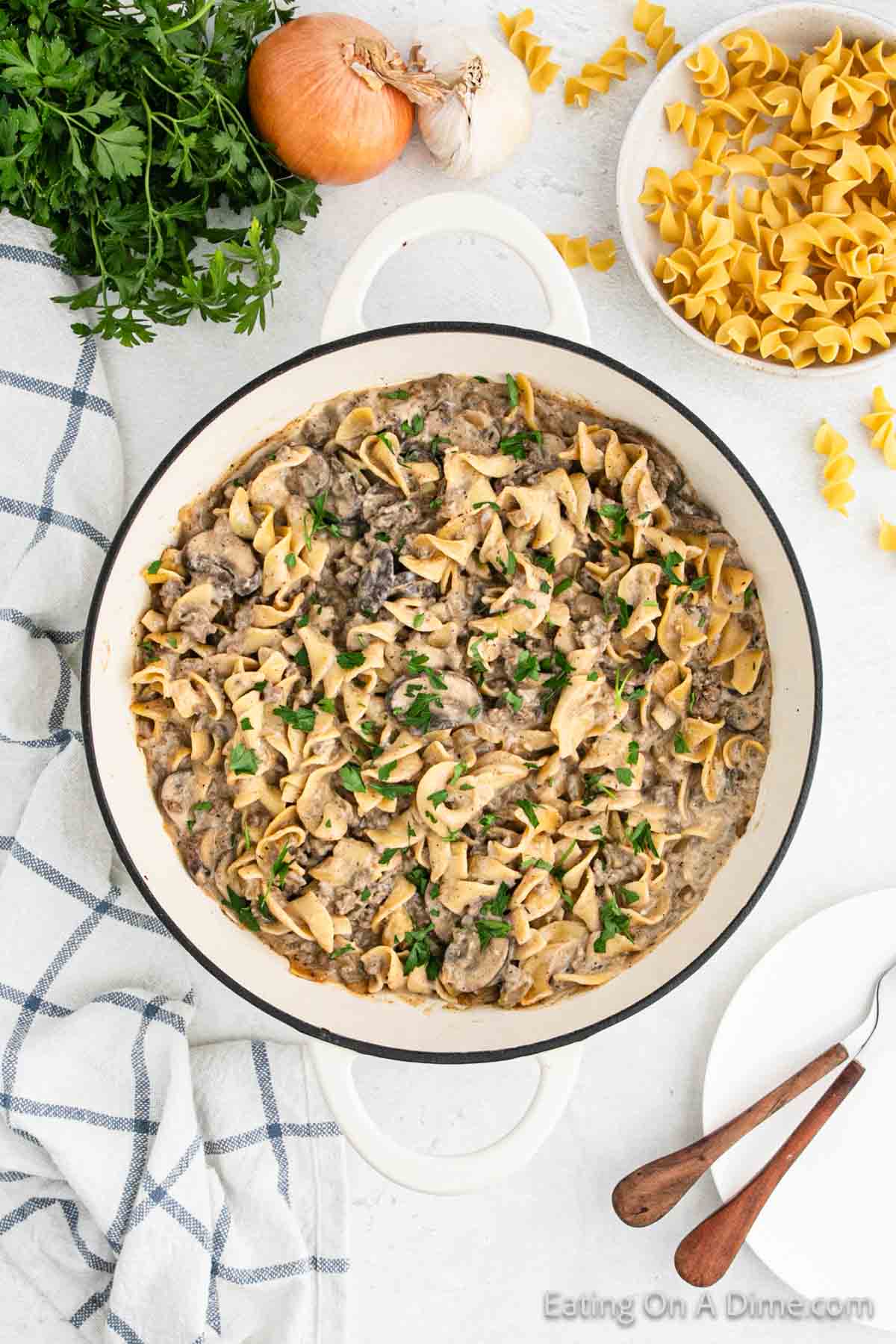 A pan of homemade beef stroganoff, reminiscent of a gourmet Hamburger Helper, features wide egg noodles mixed with tender beef strips in a creamy mushroom sauce. Fresh parsley is sprinkled on top. Surrounding the pan are ingredients like an onion, garlic, parsley, and uncooked egg noodles, with a kitchen towel and utensils nearby.