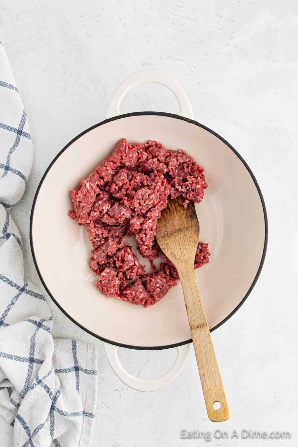 A round white pot contains chunks of raw ground beef and a wooden spoon, ready to be turned into a hearty Stroganoff. The pot is placed on a light-colored surface with a white and blue checkered cloth to the left of it, evoking comfort and home-cooked meals like Hamburger Helper.