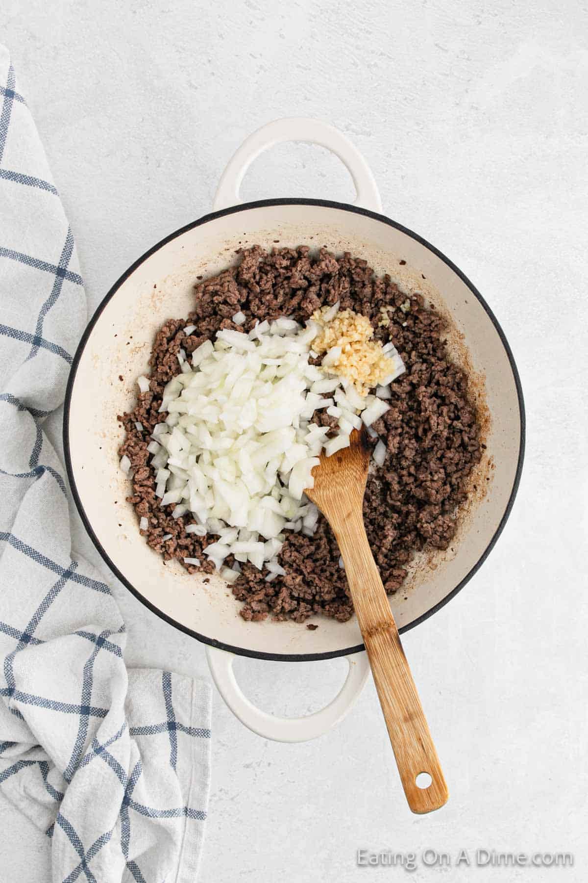 A white cast iron pot is on a light countertop, containing browned ground meat mixed with chopped white onions and minced garlic. A wooden spoon rests inside the pot, illustrating the beginnings of a savory stroganoff recipe. A blue and white checkered towel is folded next to the pot.