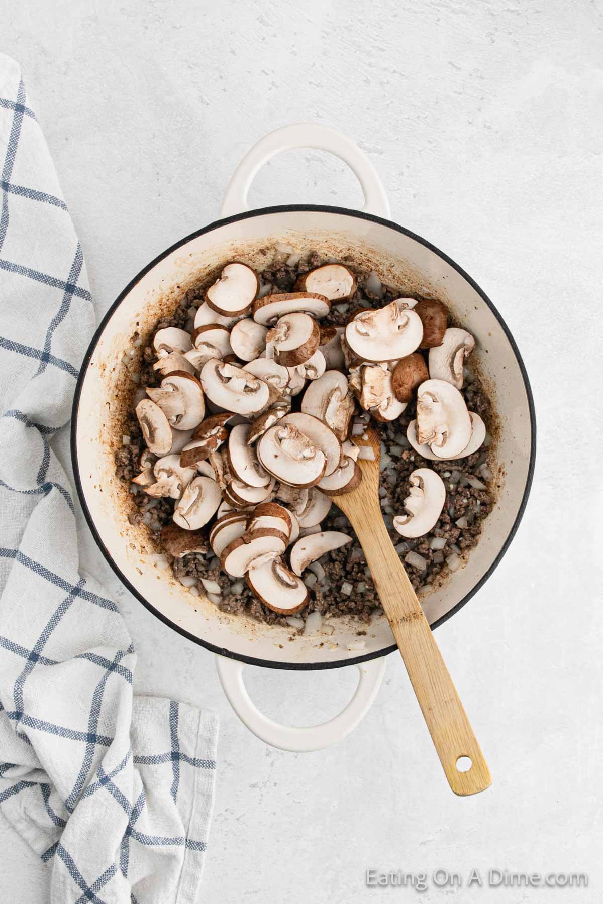 A white pot filled with sliced mushrooms being cooked with a wooden spoon stirring them, perfect for a creamy Stroganoff. The pot sits on a light countertop next to a blue and white checkered cloth.