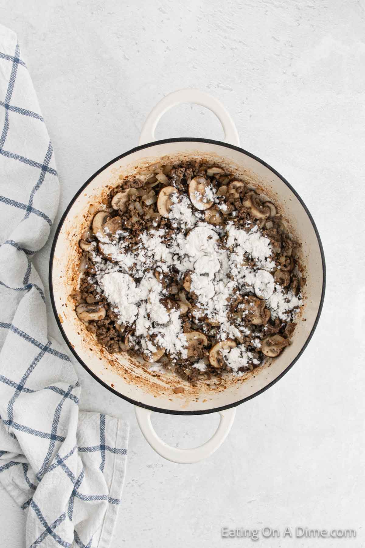 A white pot filled with sliced mushrooms and ground meat, topped with flour, reminiscent of a classic Hamburger Helper Stroganoff. The pot is placed on a light gray surface with a striped kitchen towel on the left.