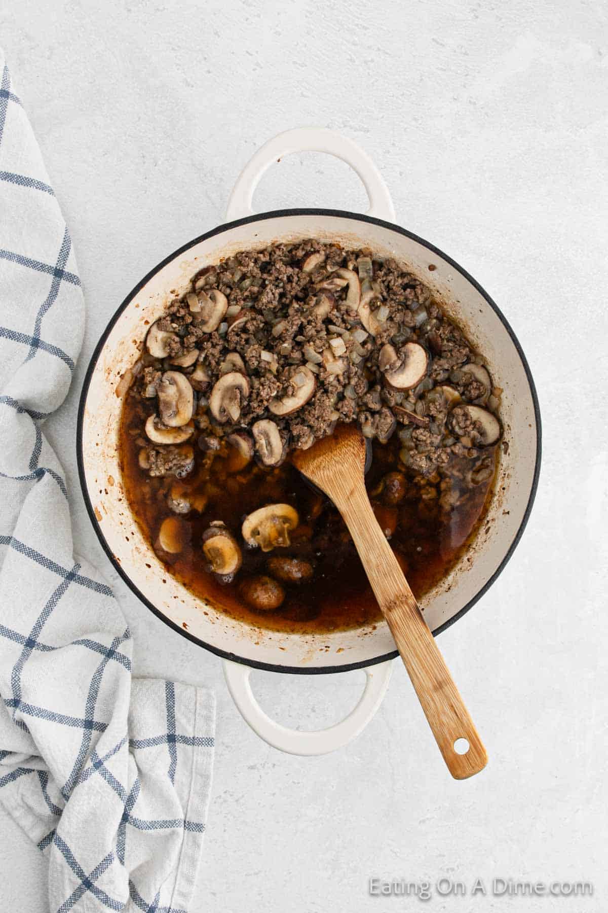 A large white pot with handles contains a delectable Hamburger Helper mixture of ground beef and sliced mushrooms in a rich brown sauce. A wooden spoon rests inside the pot, ready for serving. A white and blue checkered kitchen towel is placed to the left of the pot.