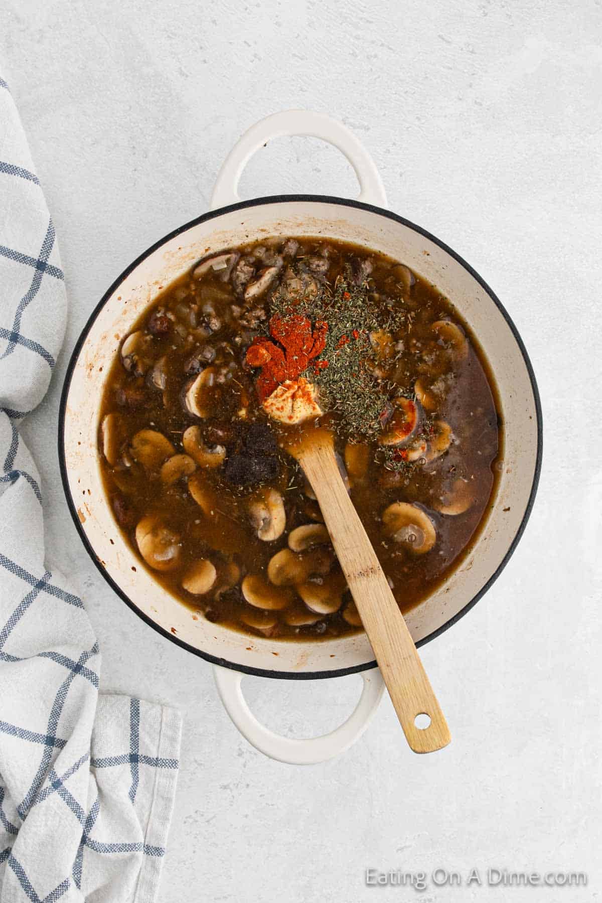 A white pot on a light surface containing a dark brown broth with sliced mushrooms, reminiscent of Hamburger Helper Stroganoff. A wooden spoon rests inside, coated in the savory broth, surrounded by a white and blue striped kitchen towel. Dried herbs and spices are visible on top of the stew. Text reads, "Eating On A Dime.com.