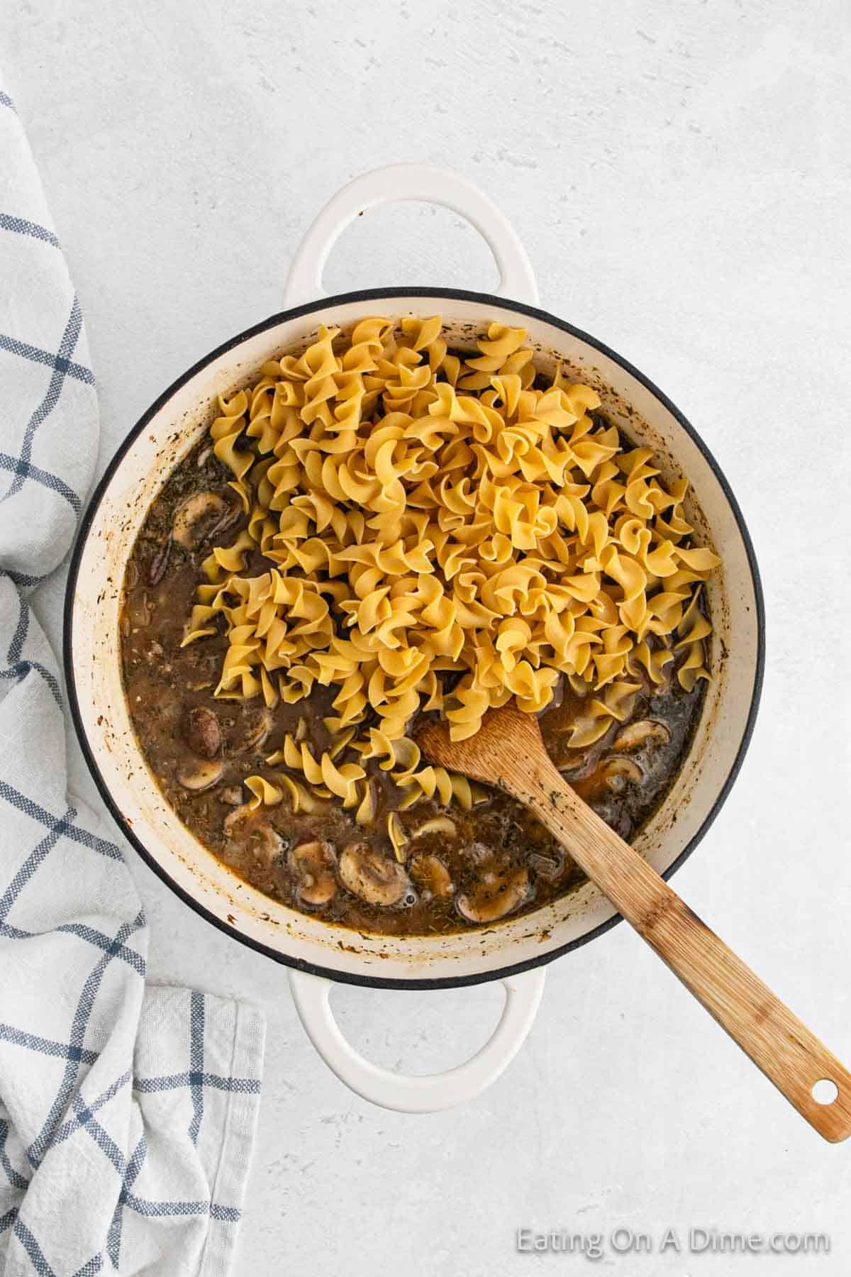 A pot filled with wide egg noodles and a dark broth with mushrooms evokes the comfort of a classic Stroganoff. A wooden spoon rests inside the pot next to the savory noodles, while a blue and white checkered cloth lies beside the pot on a white surface.