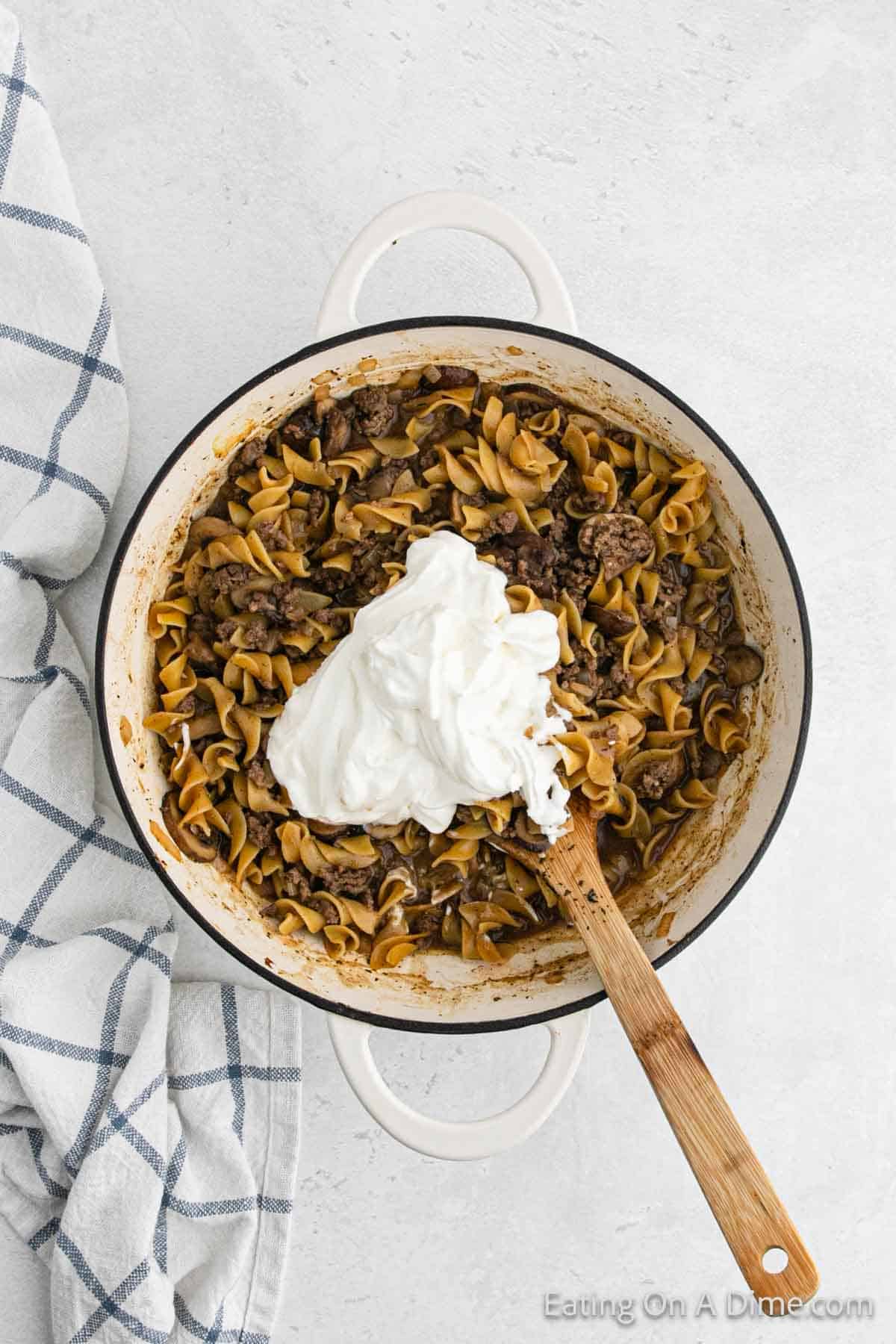 A round white pot filled with cooked noodles, ground beef, and a large dollop of sour cream sits on a light surface. A wooden spoon rests inside the pot. A white and blue checkered cloth is partially visible on the left side of the image, showcasing a classic Hamburger Helper Stroganoff meal.