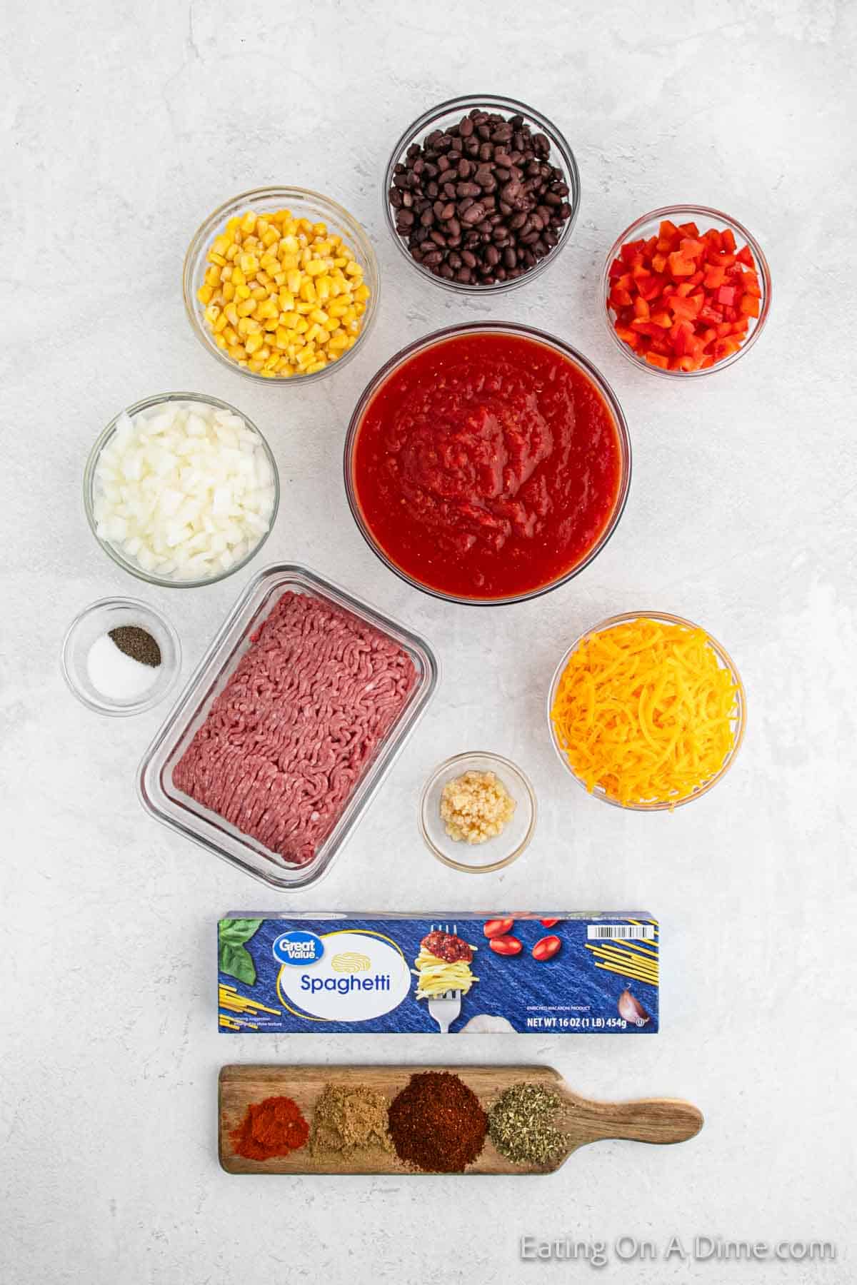 A top-down view of ingredients arranged on a white surface for Mexican Spaghetti. Includes ground meat, spaghetti, shredded cheese, diced onions, corn, black beans, bell peppers, garlic, tomato sauce, and various spices. "Eating On A Dime" is printed on the image.