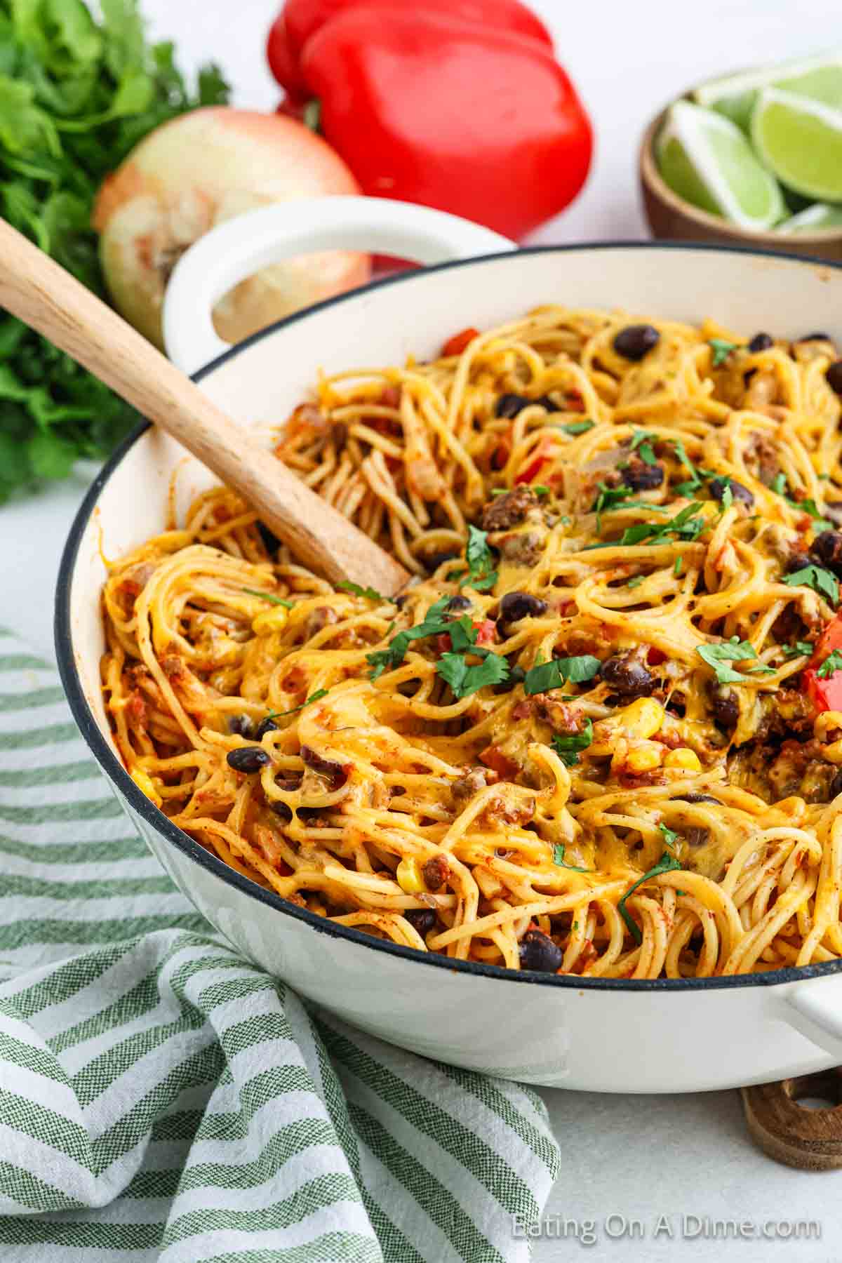 A pot of cheesy taco spaghetti, a delightful Mexican Spaghetti recipe, topped with fresh cilantro and featuring a wooden spoon for serving. The dish includes ground meat, black beans, and diced tomatoes. In the background, there are red bell peppers, an onion, lime wedges, and a bunch of cilantro. A striped cloth napkin is nearby.